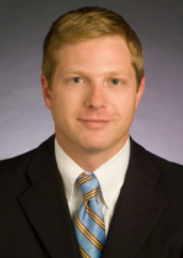 Professional headshot of a young man with short blonde hair, wearing a black suit, white shirt, and a blue striped tie, posing against a gray background.