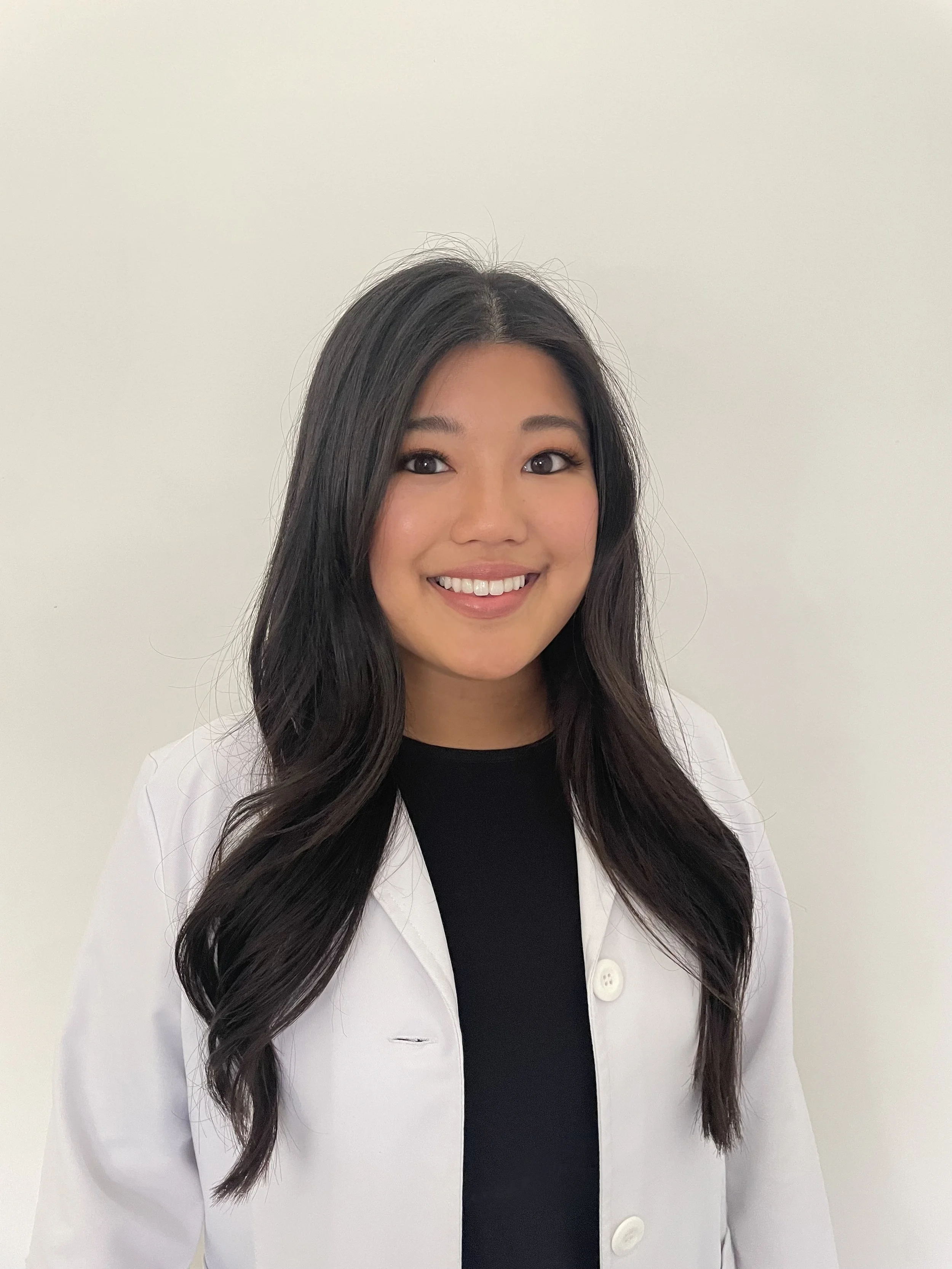 A woman with long dark hair, wearing a white coat over a black top, smiling against a plain white background.