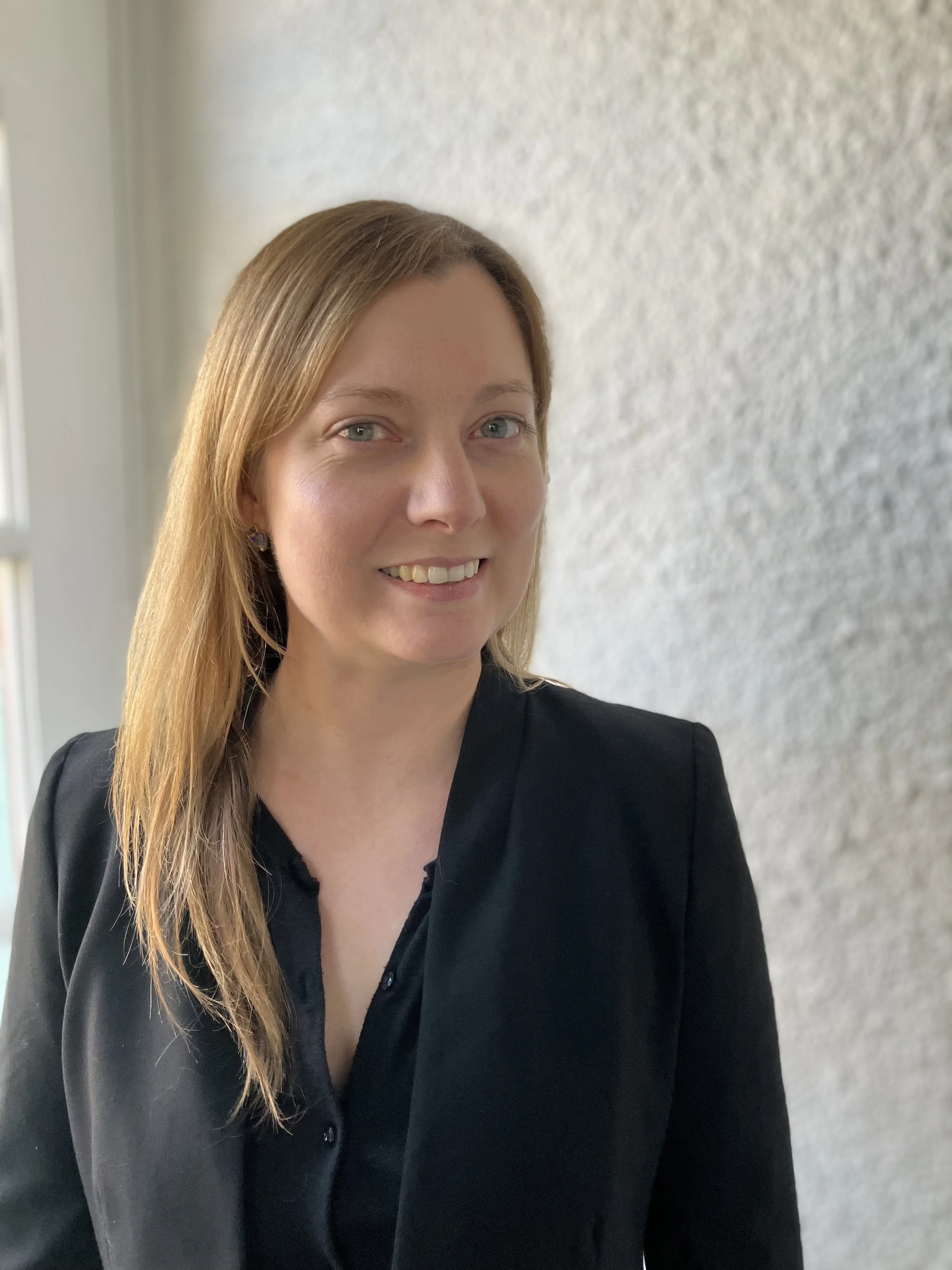 Professional woman with red hair and glasses, wearing a white coat, outdoors with greenery and sunlight in the background.