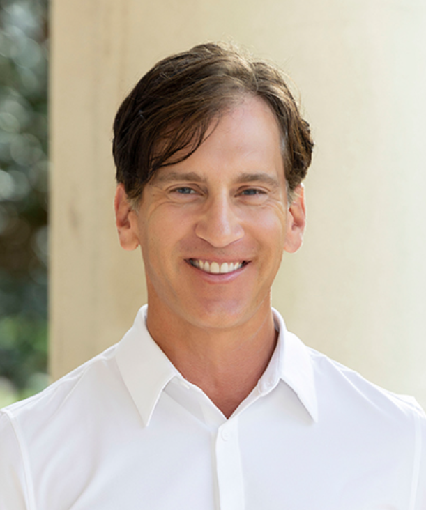 Portrait of a smiling man with brown hair wearing a white shirt, standing outdoors near a column with blurred greenery in the background.