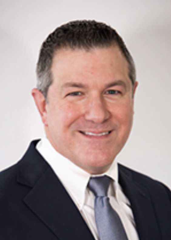 A smiling man in a dark suit, white shirt, and light gray tie against a plain light background.