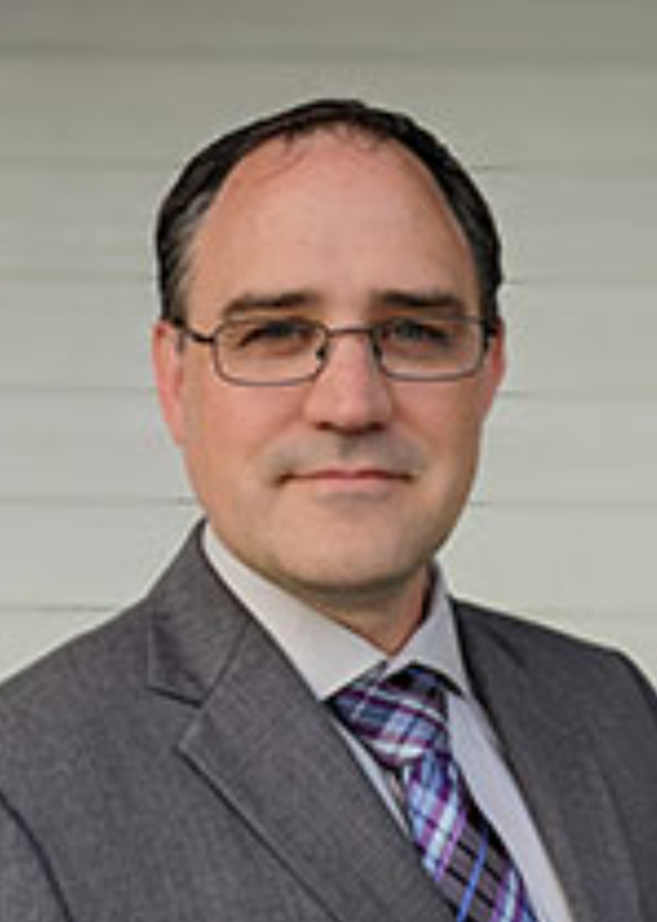 A man wearing glasses in a suit and tie, posing outdoors in front of a light-colored wall.