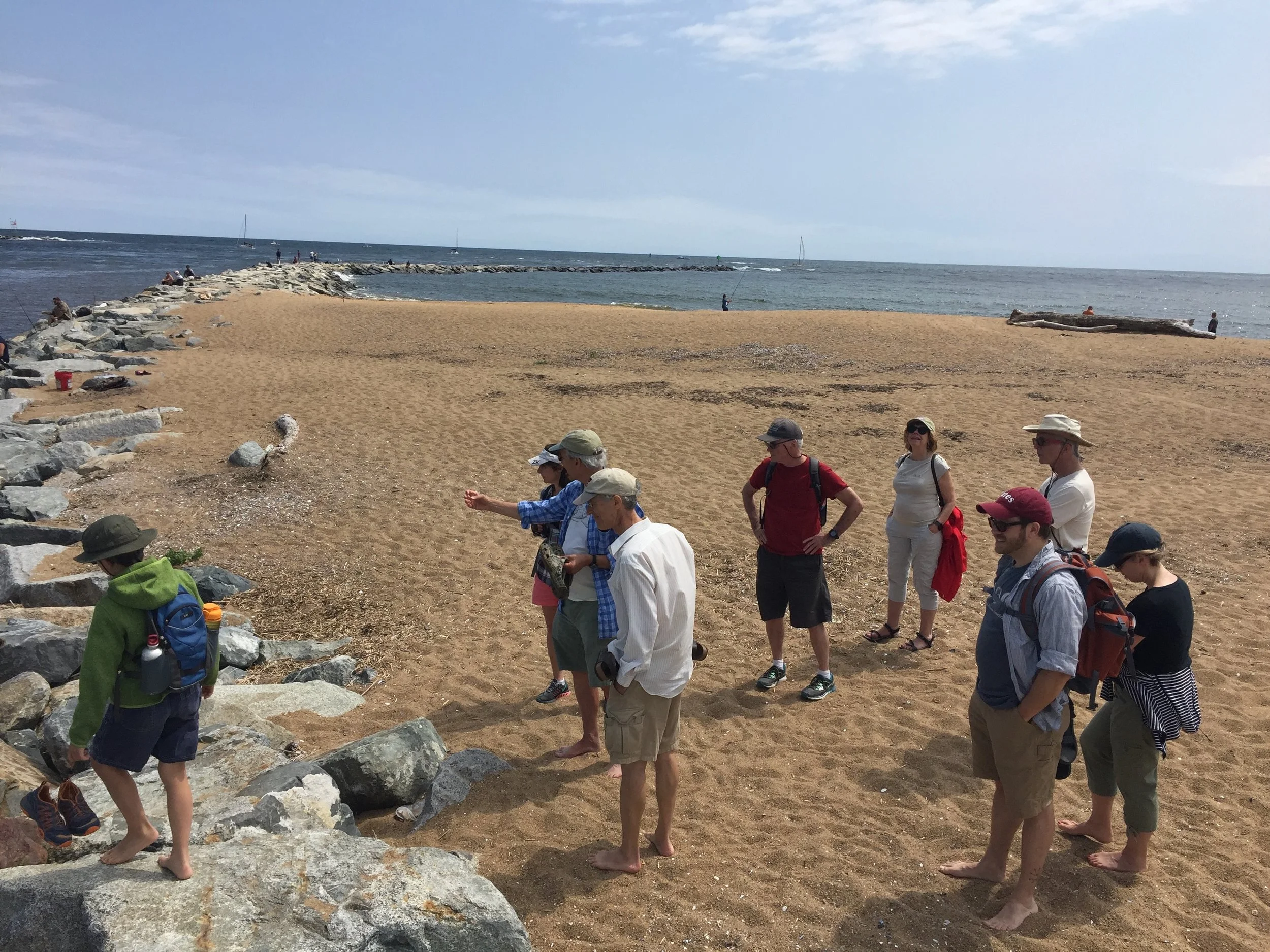 Group of nine people exploring a sandy beach near rocks with the ocean in the background, some carrying backpacks and wearing hats.
