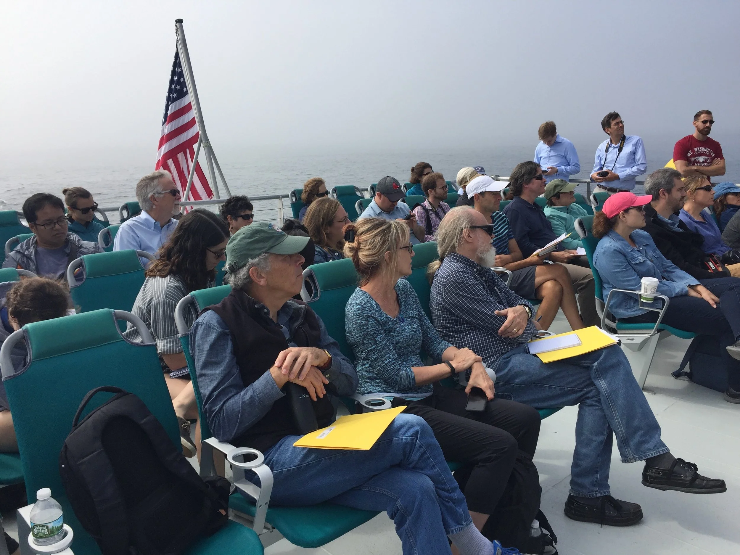 A group of people seated on a boat or ferry, attending a presentation or speech. An American flag is visible on the boat, and the background shows foggy water.