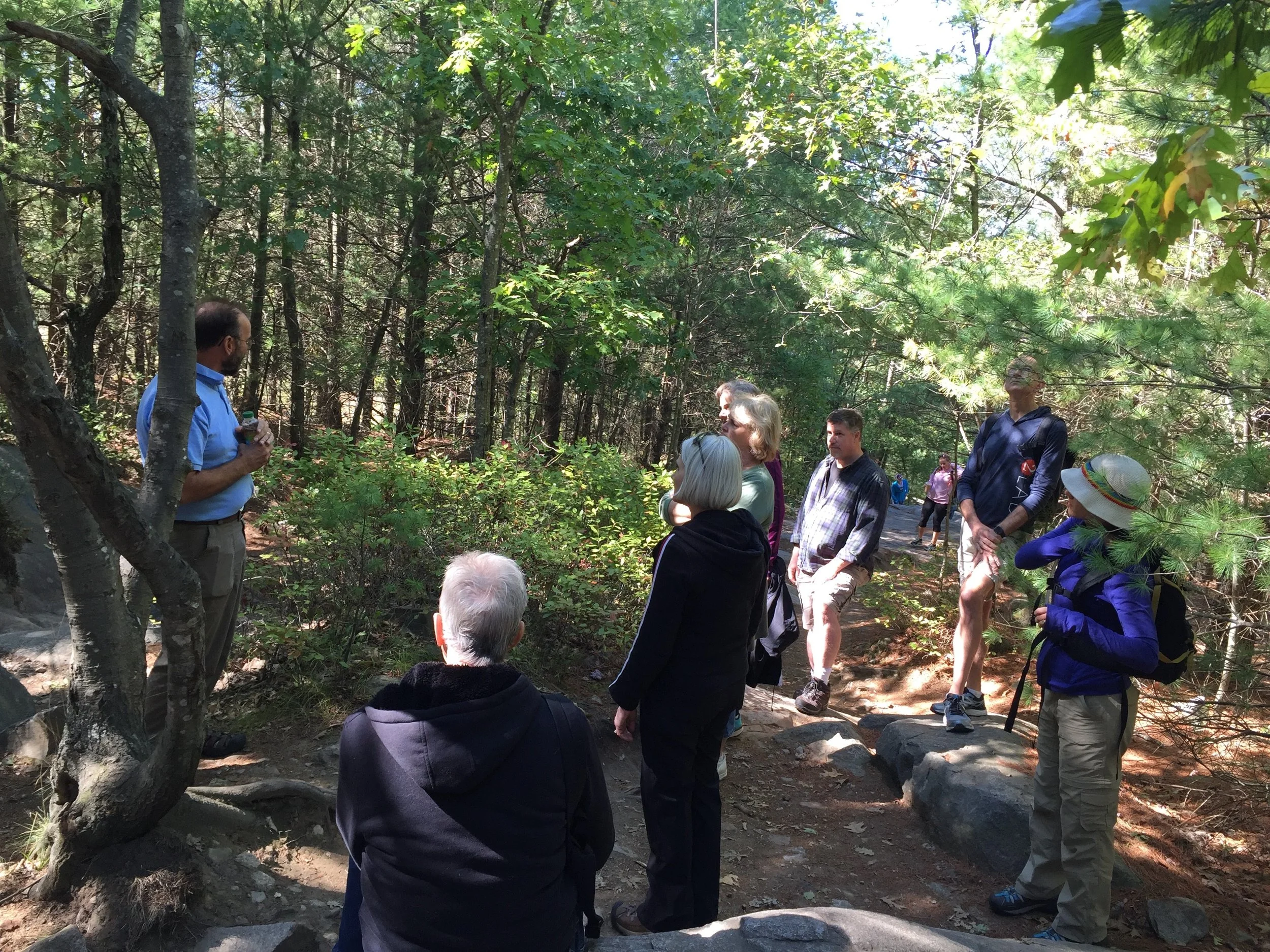 Group of people gathered in a wooded forest area, listening to a man in a blue shirt giving a talk or presentation.