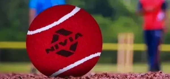 Close-up of a red cricket ball on the ground with a blurred sports field and two players in the background.