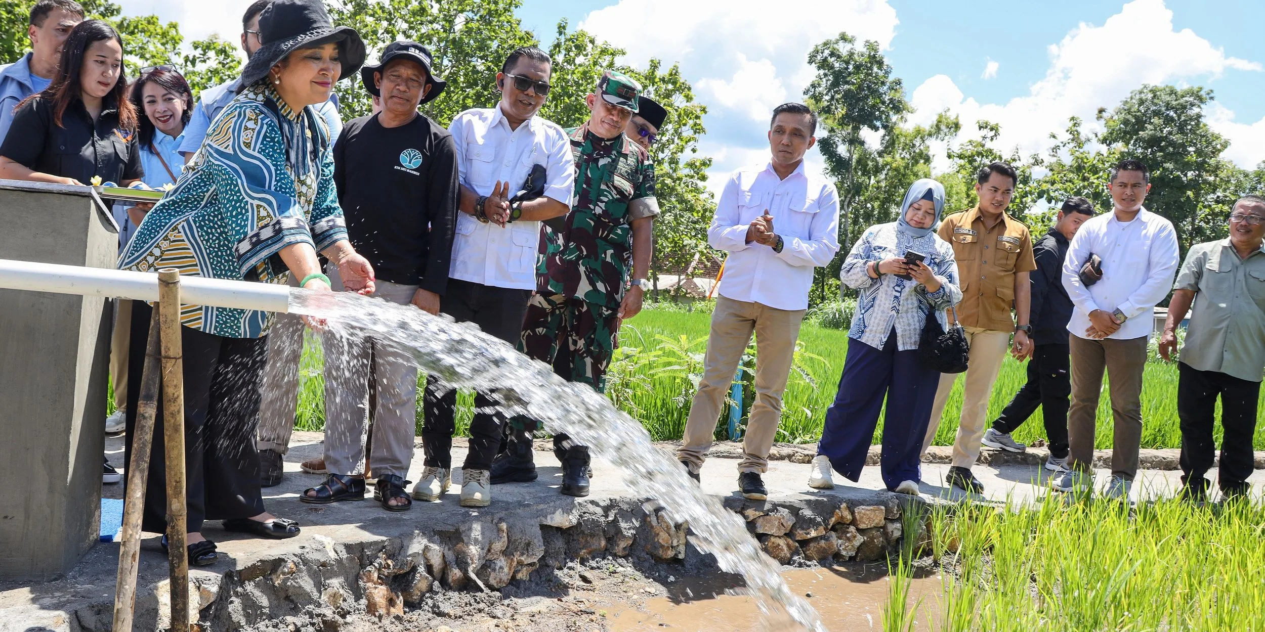 A group of people standing outdoors near a rice paddy, with a woman in a large hat pouring water from a pipe, and others watching and taking photos during the daytime.