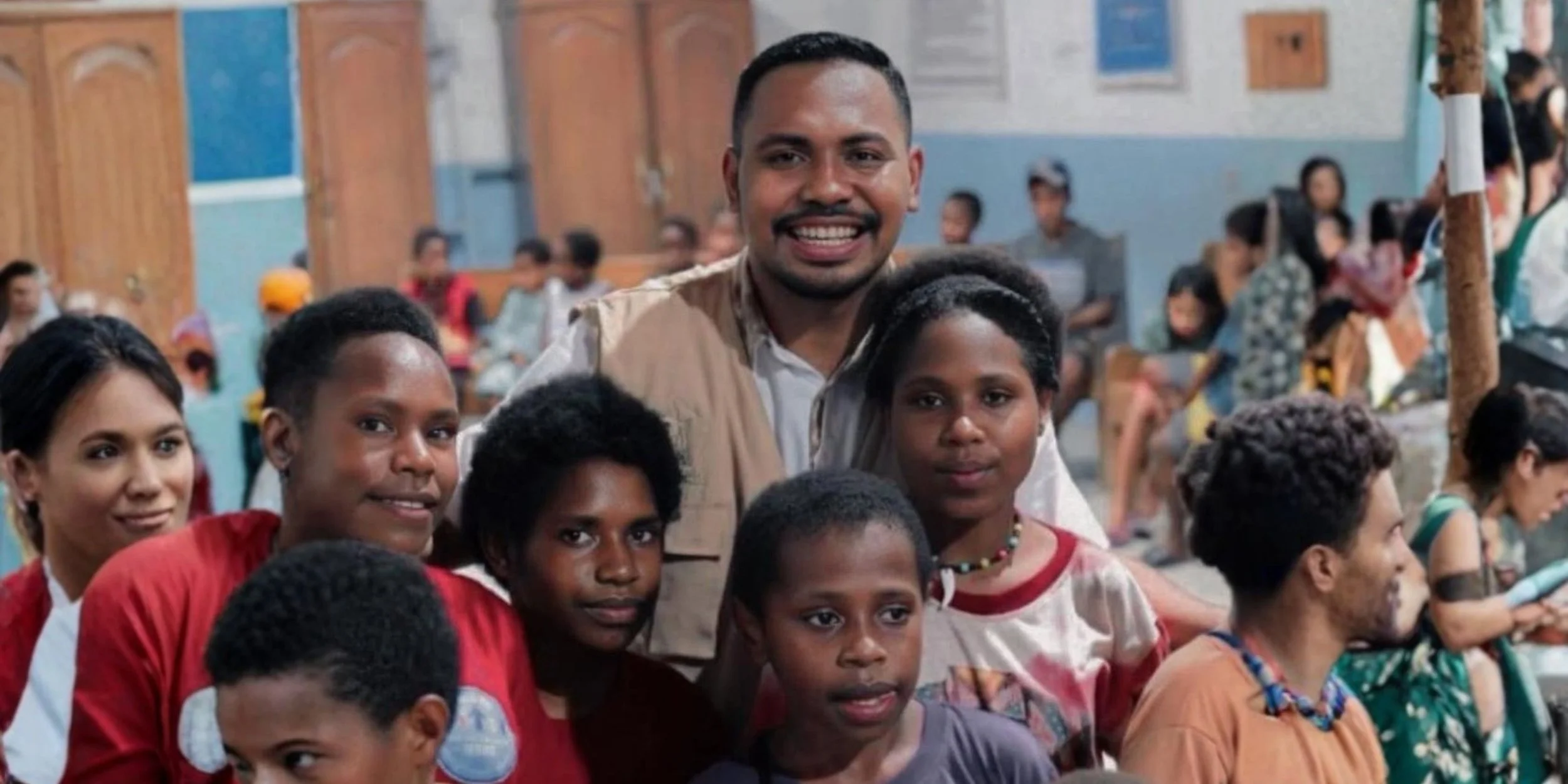 A diverse group of children and an adult man, smiling and posing together in a crowded indoor setting, possibly a school or community center.