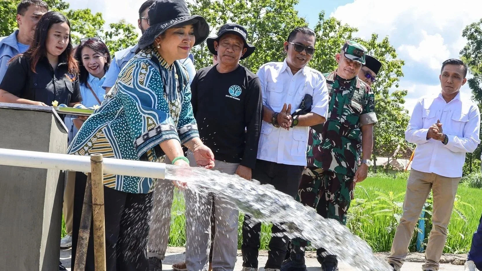 A group of people standing outdoors near a rice paddy, with a woman in a large hat pouring water from a pipe, and others watching and taking photos during the daytime.
