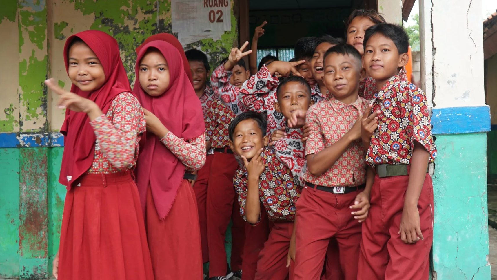 Group of children in colorful uniforms, smiling and posing outside a building, some making peace signs.