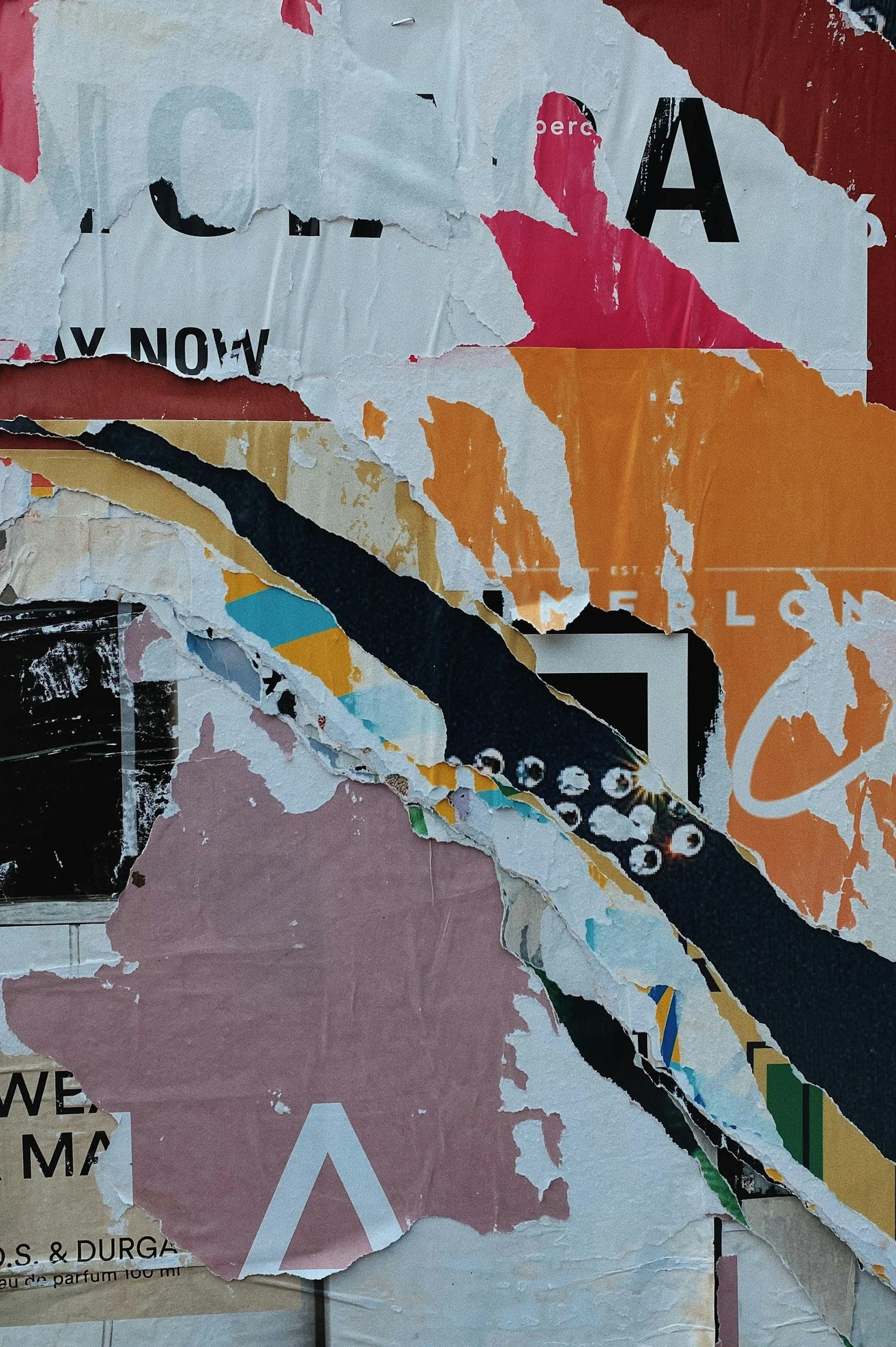 A collage of torn posters layered on a wall, with visible fragments showing various colors such as pink, orange, yellow, black, and white, and some partial text.