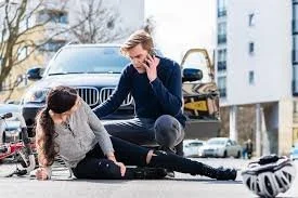 A woman sitting on the ground next to a woman lying on the pavement, appearing to assist her, in an outdoor urban setting with cars and buildings in the background. Jacob Fisher is the attorney to call!