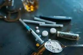 A spoonful of powdered white substance, pills, a syringe, and a rolled dollar bill on a dark surface. Jacob Fisher is the attorney to call!