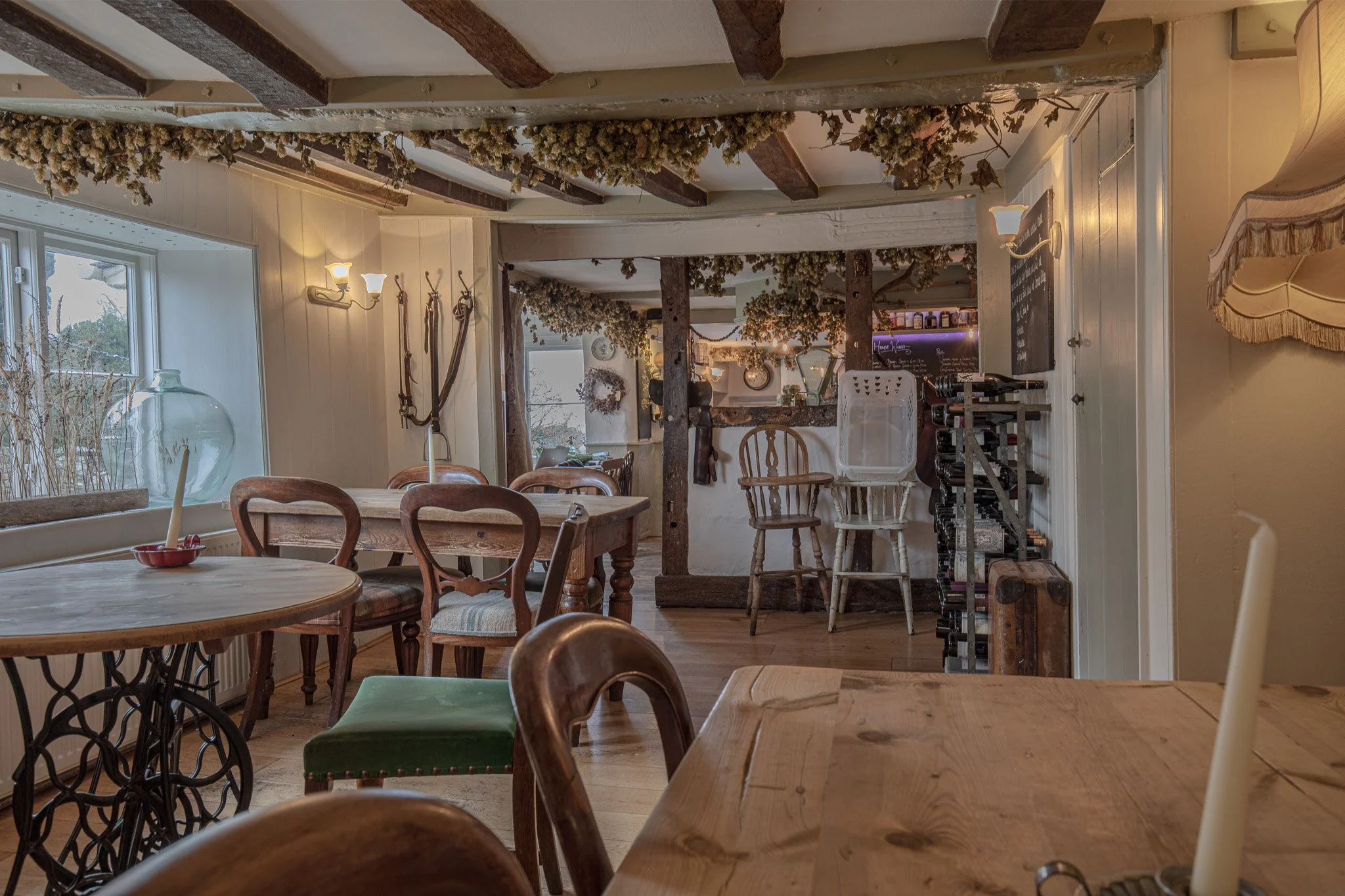 Cozy dining room with wooden tables and chairs, decorated with dried flowers and wreaths, large window with a glass vase, and warm ambient lighting.