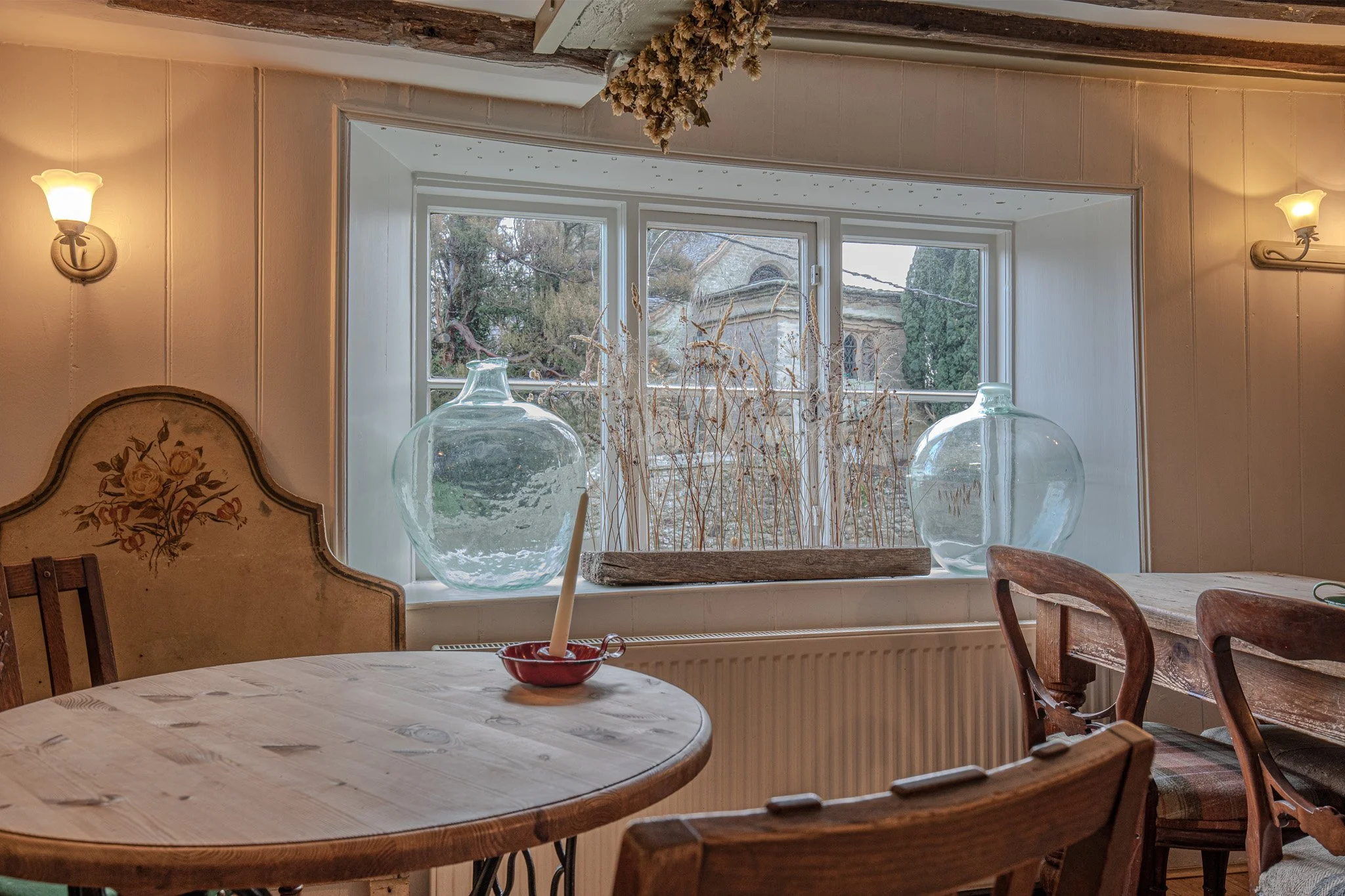 Cozy dining room with a round wooden table, vintage chairs, a large window with decorative glass jars, and dried plants on the window sill. Warm lighting from wall sconces.