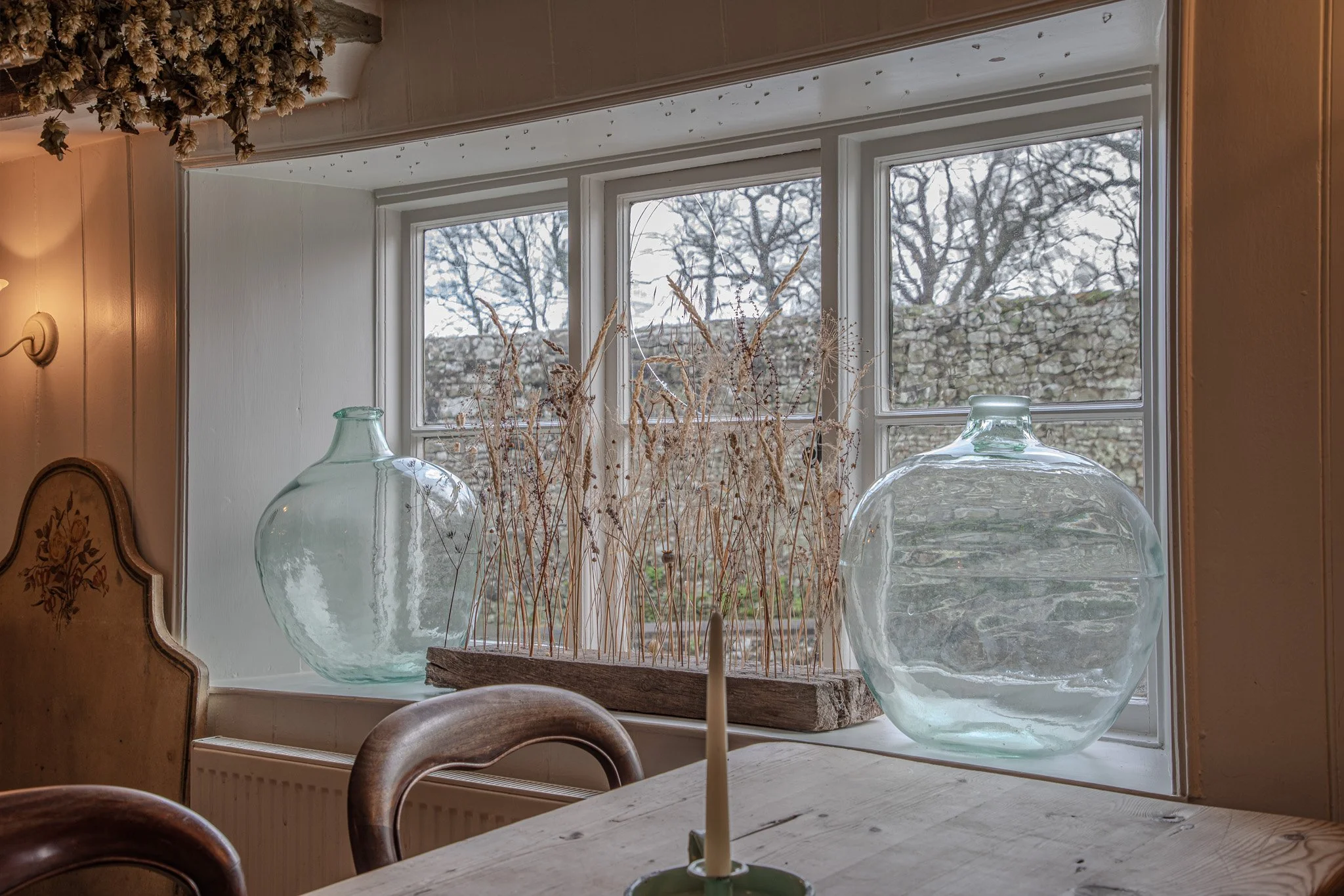 Decorative glass vases, dried plants, and a candle on a table near a window with a stone wall and trees outside.