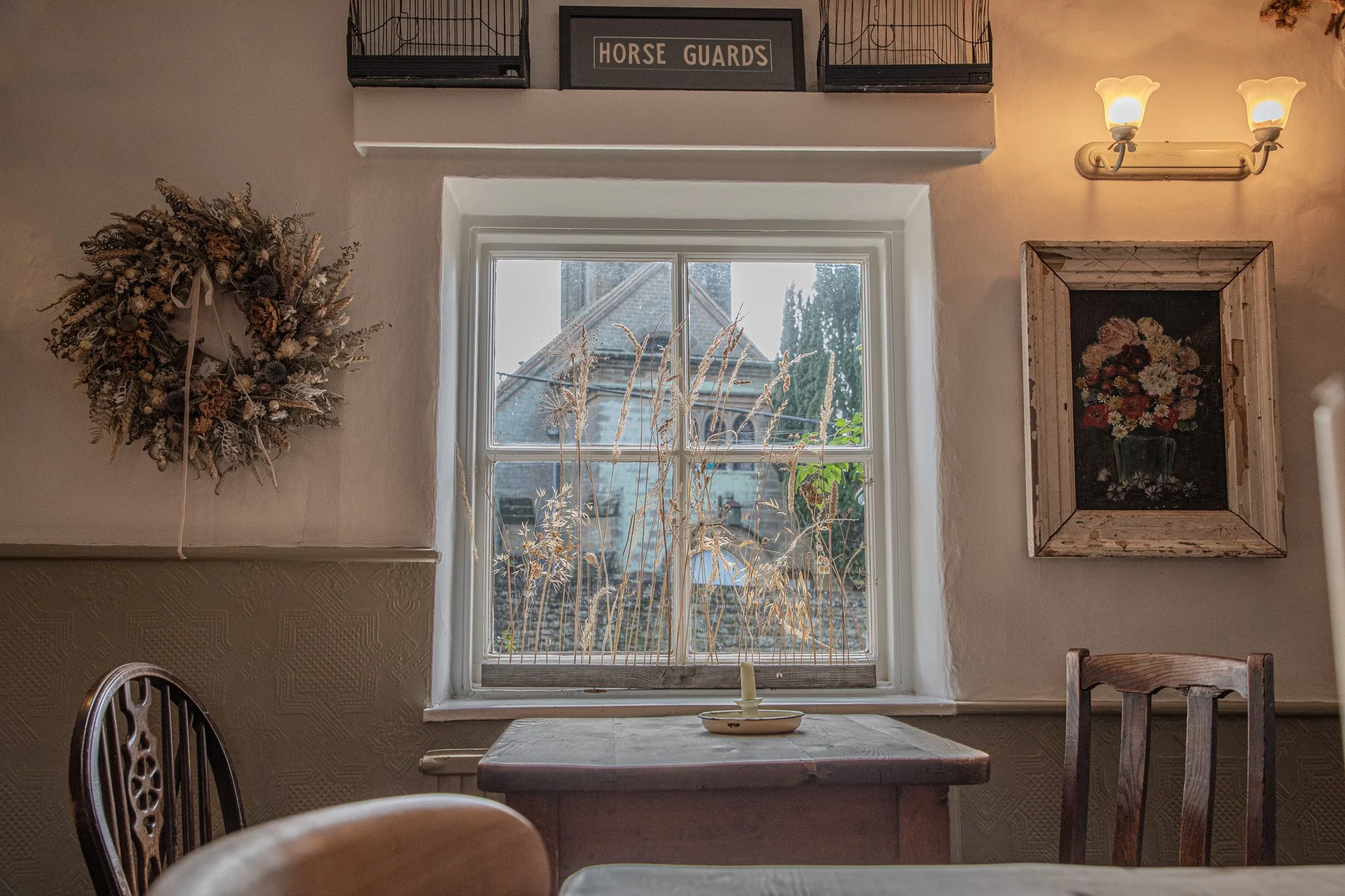 Interior of a cozy room with a wooden table and chairs, a window with dried grass plants outside, a floral wreath on the wall, a framed flower painting, and a sign labeled 'Horse Guards' on a shelf above the window. There is a double-bulb wall light 