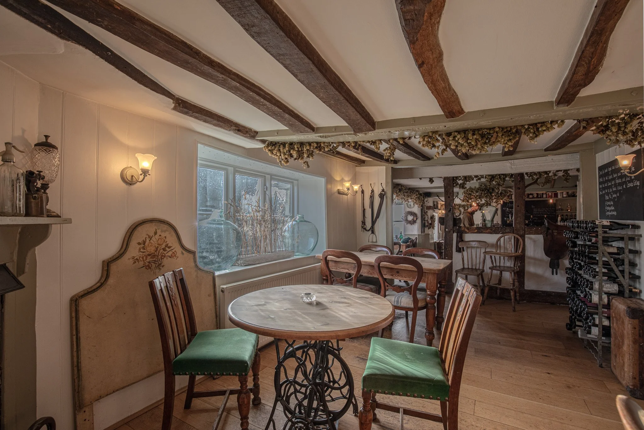 Cozy indoor dining area with wooden beams on ceiling, vintage chairs with green cushions, a round marble-topped table, and decorative glass jars on windowsill. Soft lighting and rustic decor create warm atmosphere.