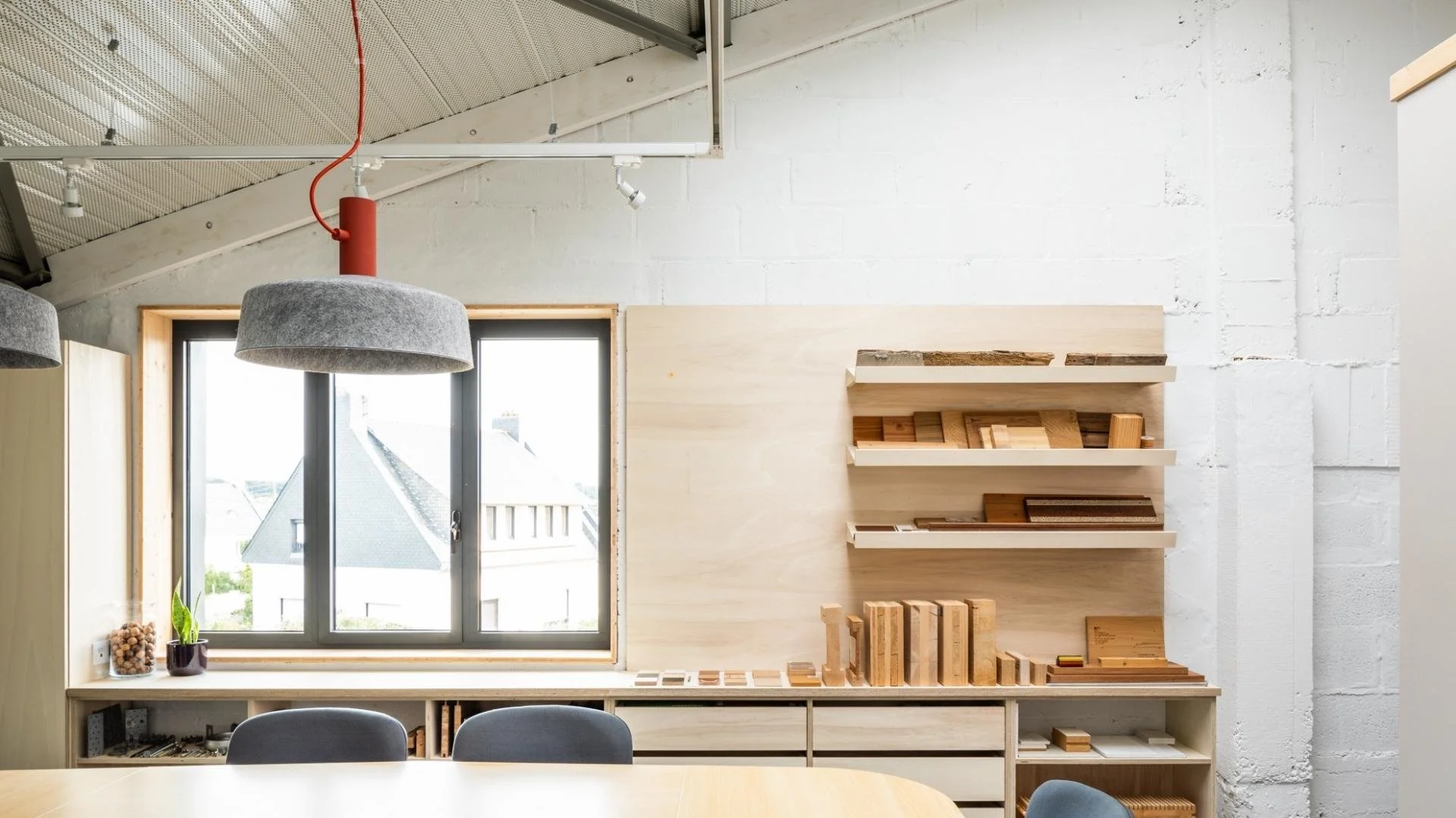 Aménagement de bureau style industriel avec mobilier en bois clair, étagères murales et grandes fenêtre offrant une vue urbaine