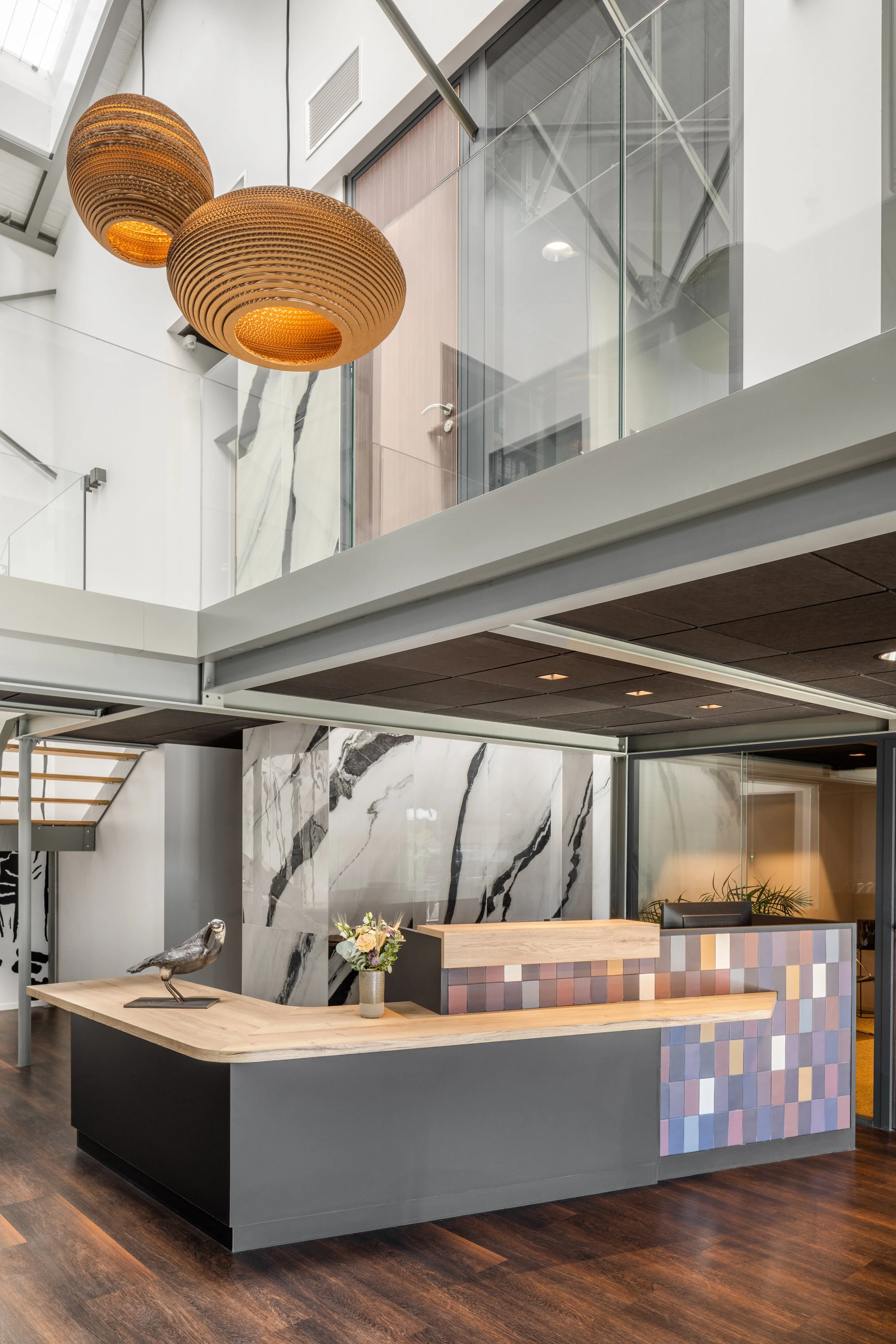 Un intérieur moderne avec un comptoir en bois, une décoration en mosaïque de céramique aux couleurs pastel, un vase avec des fleurs, une statue d'oiseau, et des lampes suspendues sphériques en bois.