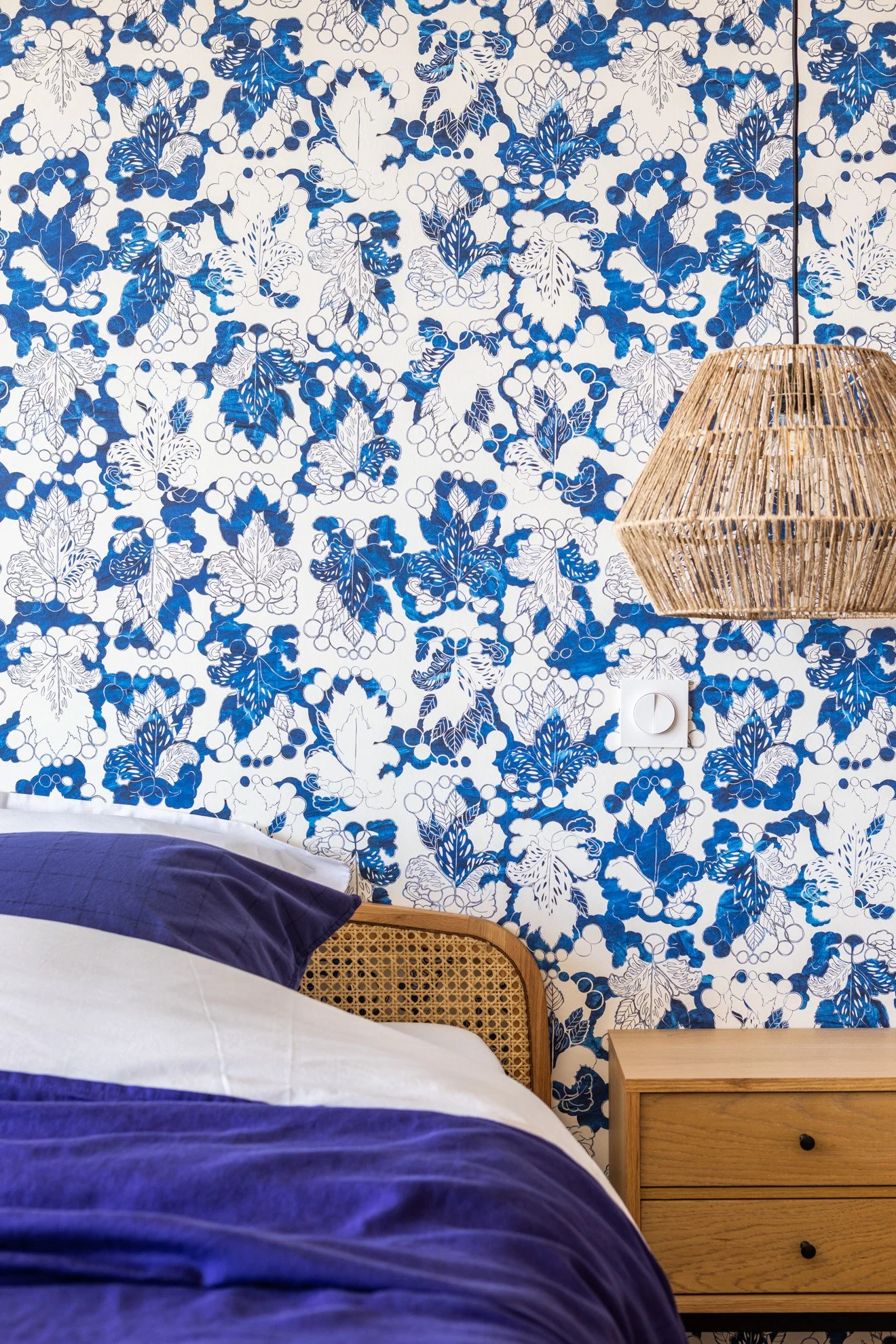 Un mur recouvert d'un papier peint à motif floral bleu et blanc, avec une lampe en rotin suspendue, une prise électrique blanche et un meuble en bois clair à tiroirs. Une partie d'un lit avec une couverture blanche et un oreiller violet est visible en bas de l'image.