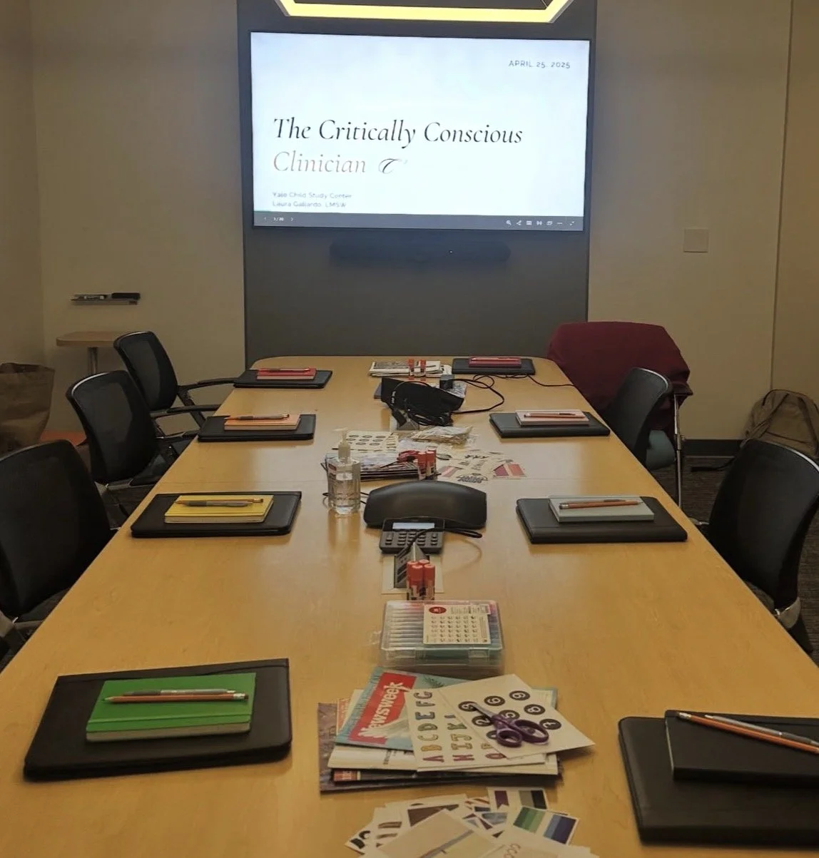 Empty conference room with a long table set for a meeting, notebooks and pens at each seat, a large screen displaying a presentation titled 'The Critically Conscious Clinician' on April 25, 2025, and various office supplies and materials on the table.