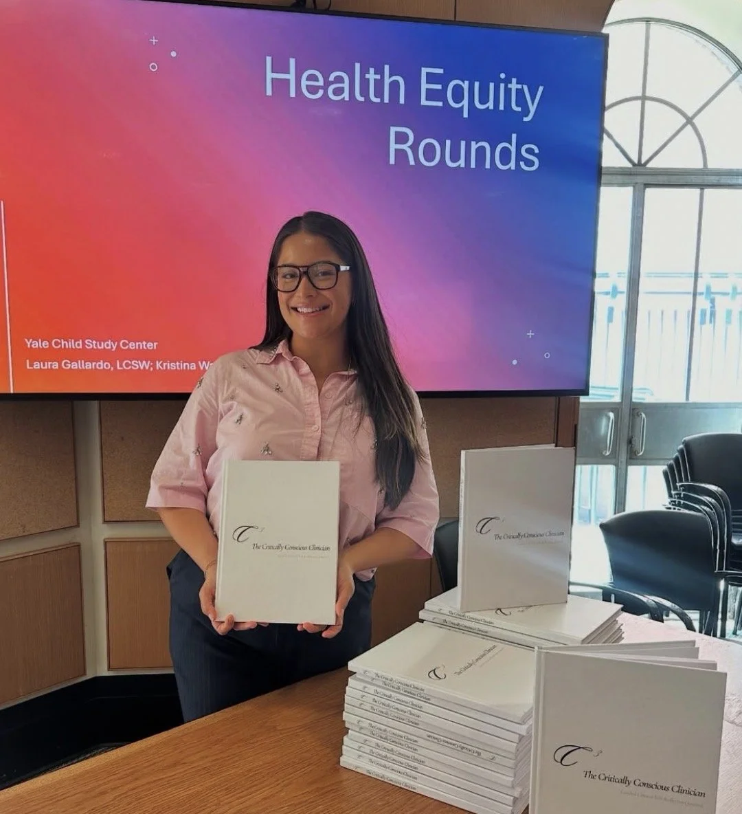 A woman with long black hair, glasses, and a light pink shirt is holding a book titled "The Critically Conscious Clinician" and smiling at the camera. Behind her is a large screen displaying "Health Equity Rounds" and a table with more copies of the same book.