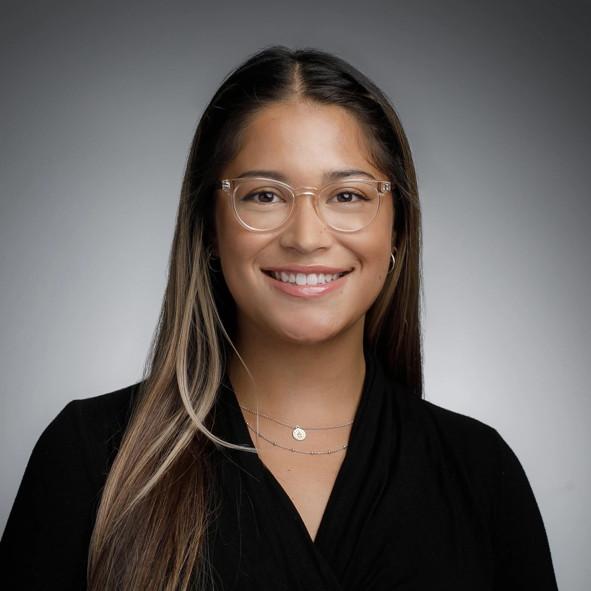 A young woman with long brown hair, glasses, and wearing a black top, smiling against a gray background.