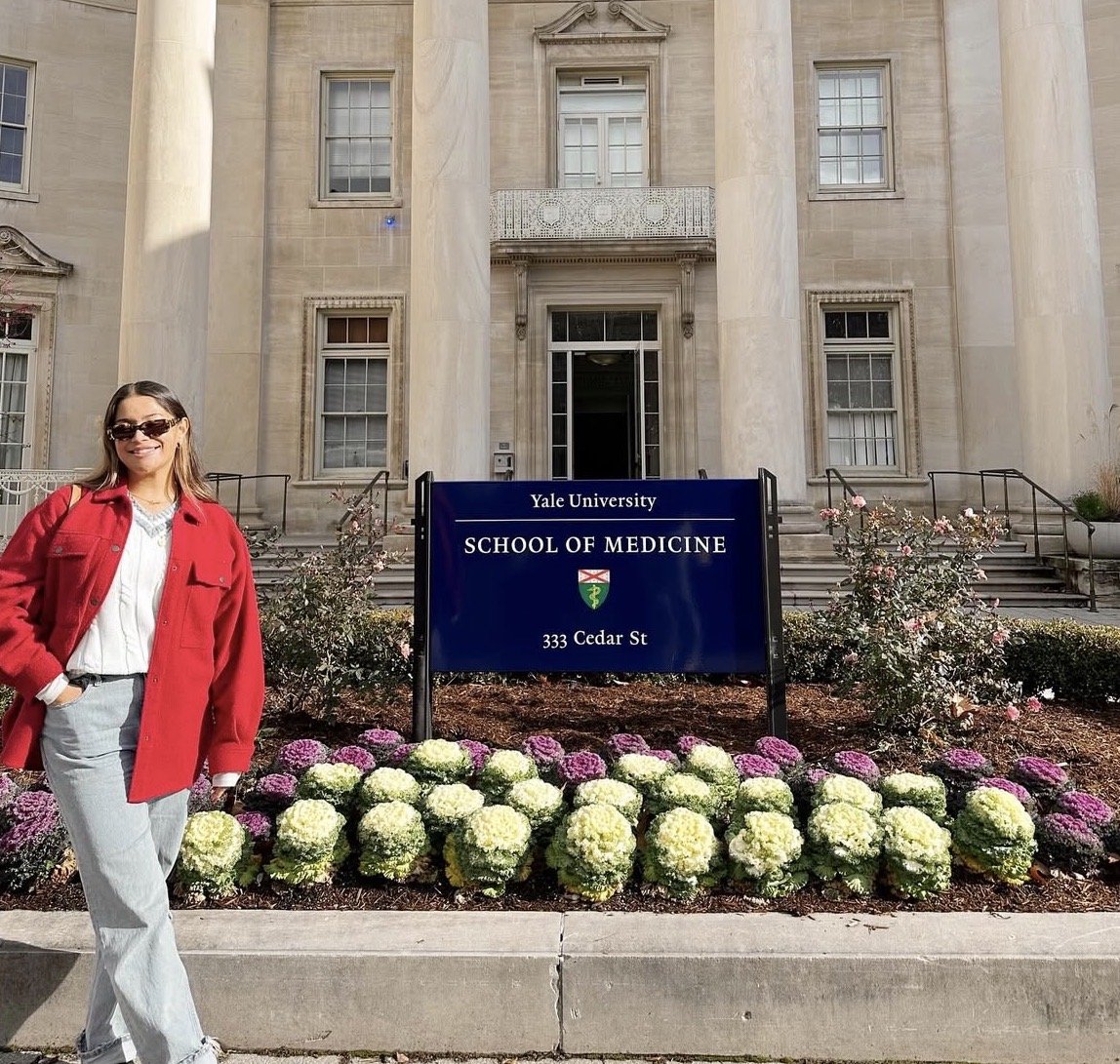 A woman wearing a red jacket and sunglasses standing in front of Yale University's School of Medicine building at 333 Cedar St, with a sign, flowers, and the building's entrance in the background.