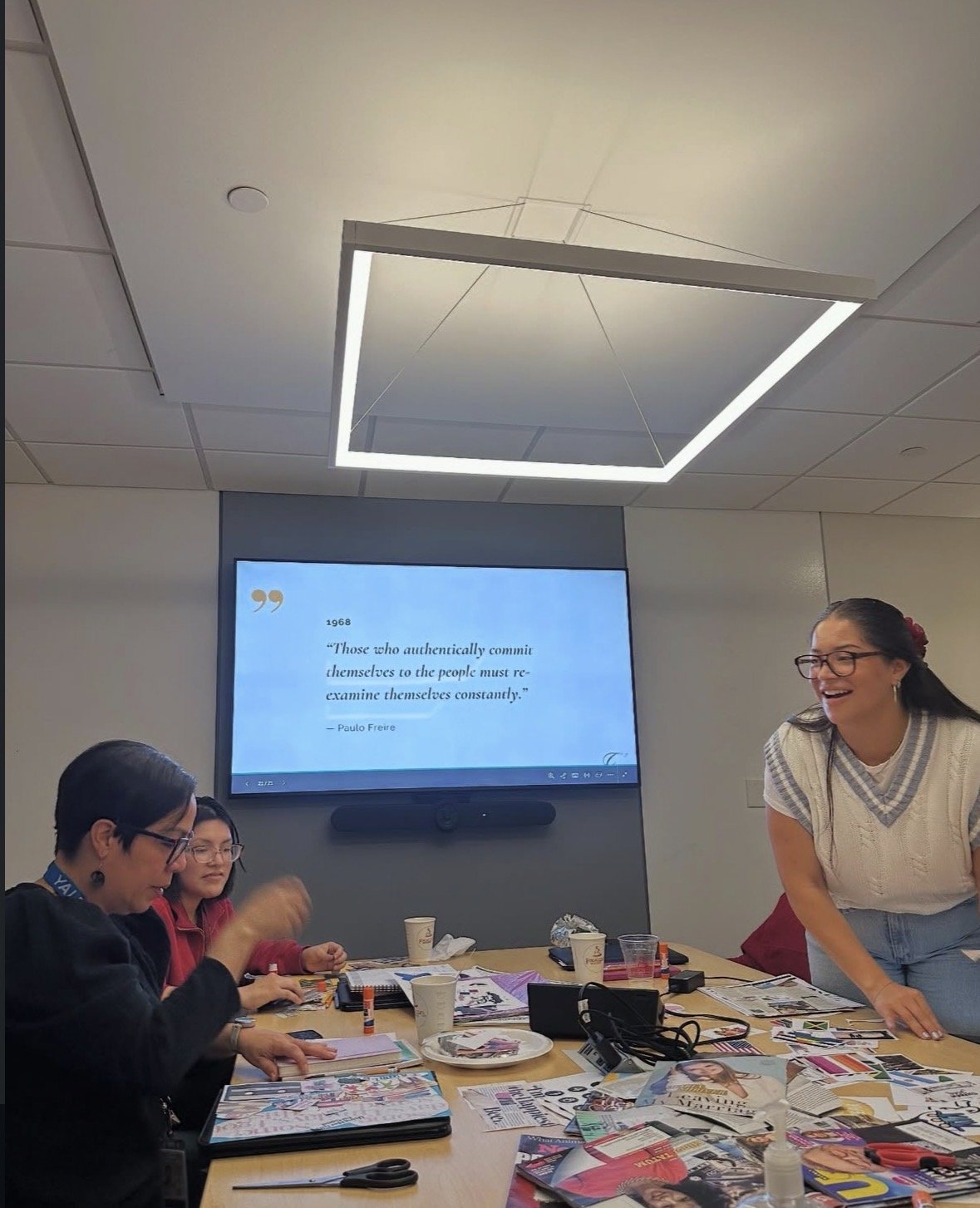 A group of women in a meeting room, some seated at a table with papers and magazines, and one woman standing and smiling. A large screen on the wall displays a quote from Paulo Freire about self-criticism.