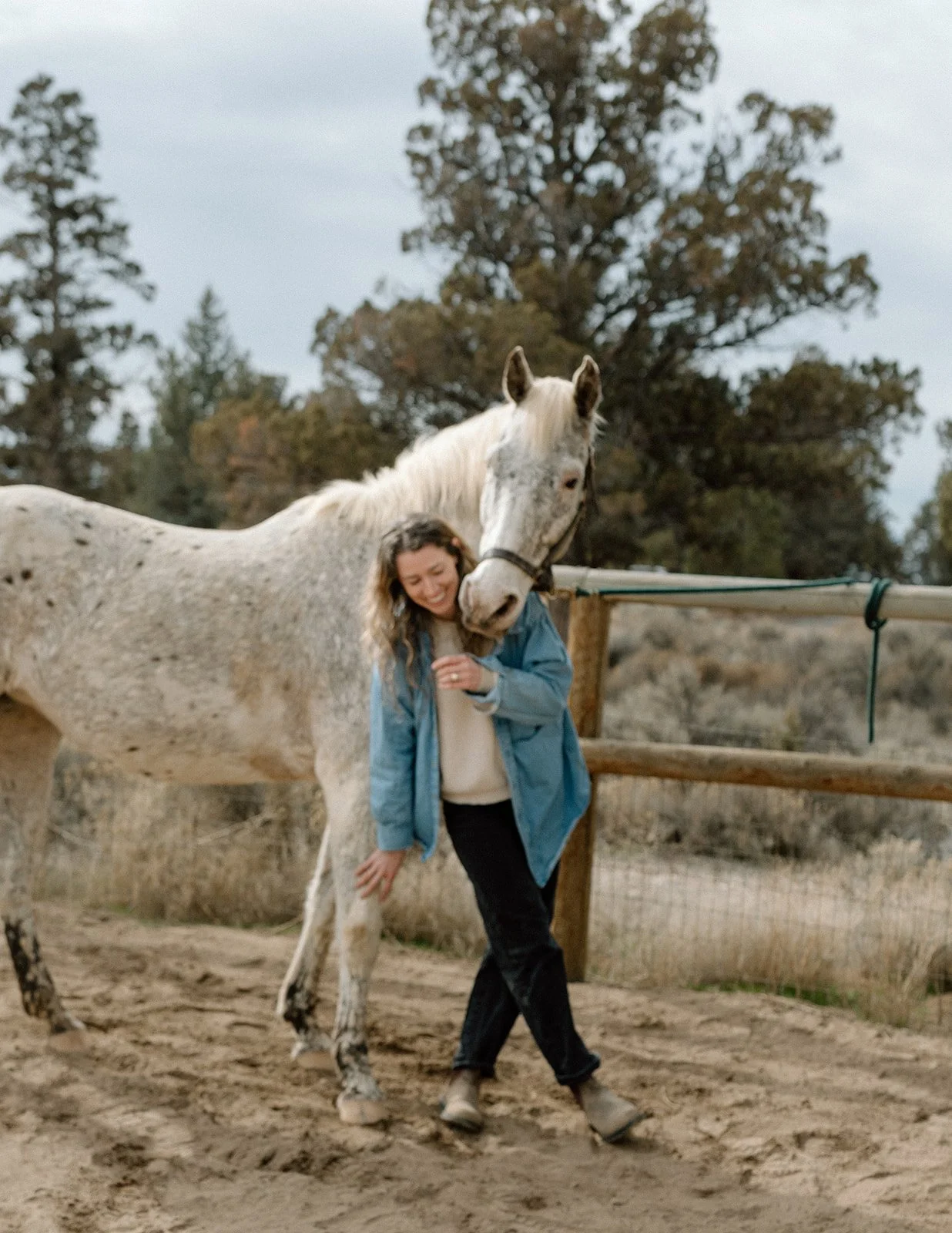 A woman, Kylen, in a blue jacket smiling and hugging Reebs, a white horse with gray spots, standing on a dirt ground near a wooden fence in an outdoor rural setting with trees in the background.
