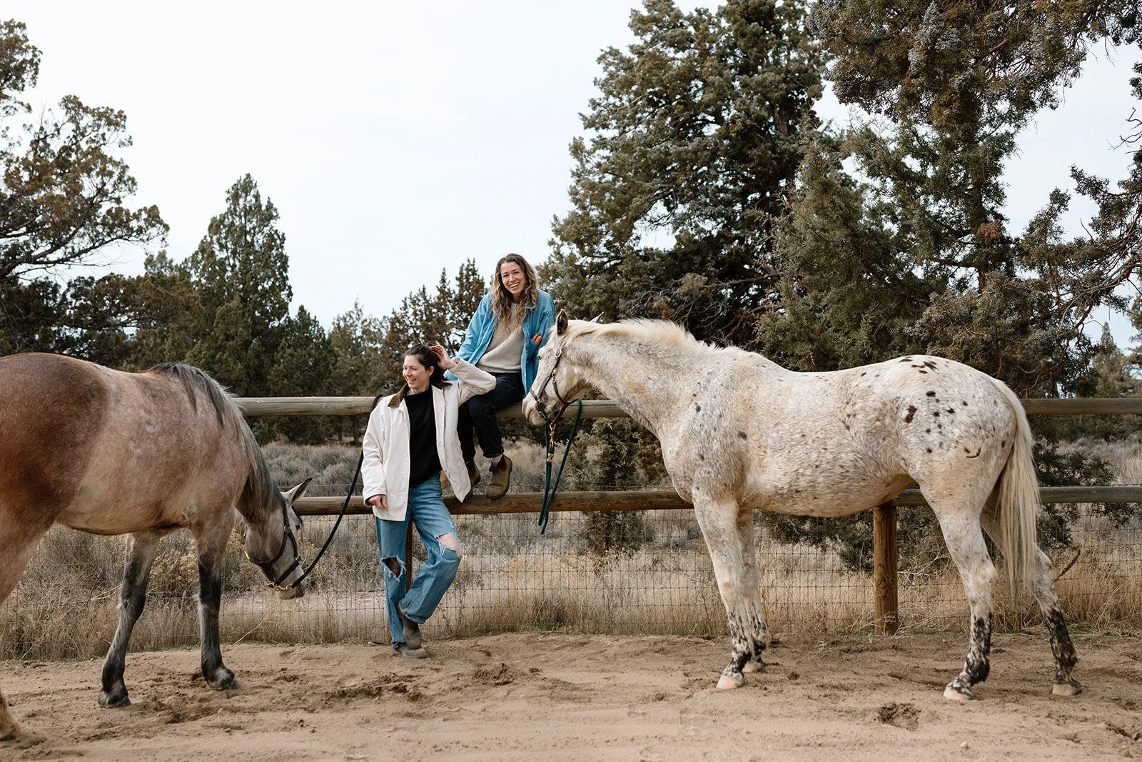 Two women, Robin and Kylen, and their two horses, Essie and Reebs, at a fenced outdoor area, with trees in the background.