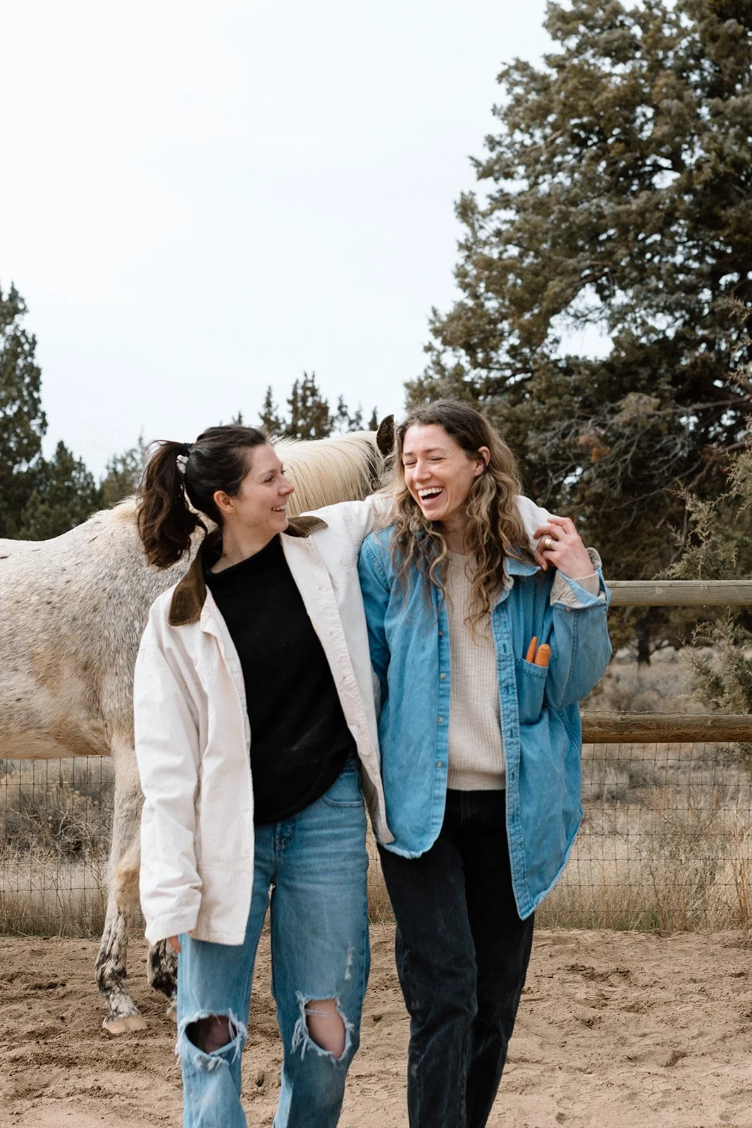 Two women, Robin and Kylen, laughing and smiling outdoors, standing close to each other, with a horse behind them, in a rustic setting with trees and wooden fencing.