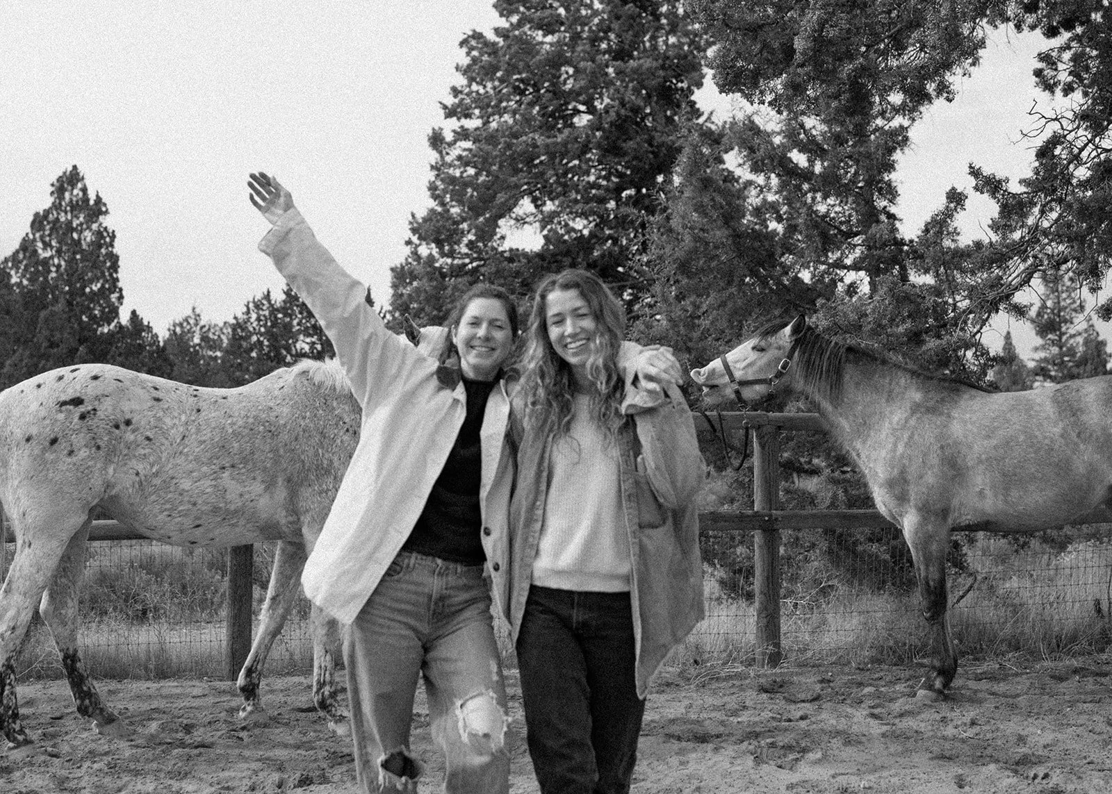Two women, Robin and Kylen, standing in front of a fence with horses behind them, smiling, one with arm raised in a friendly gesture, outdoor setting with trees.