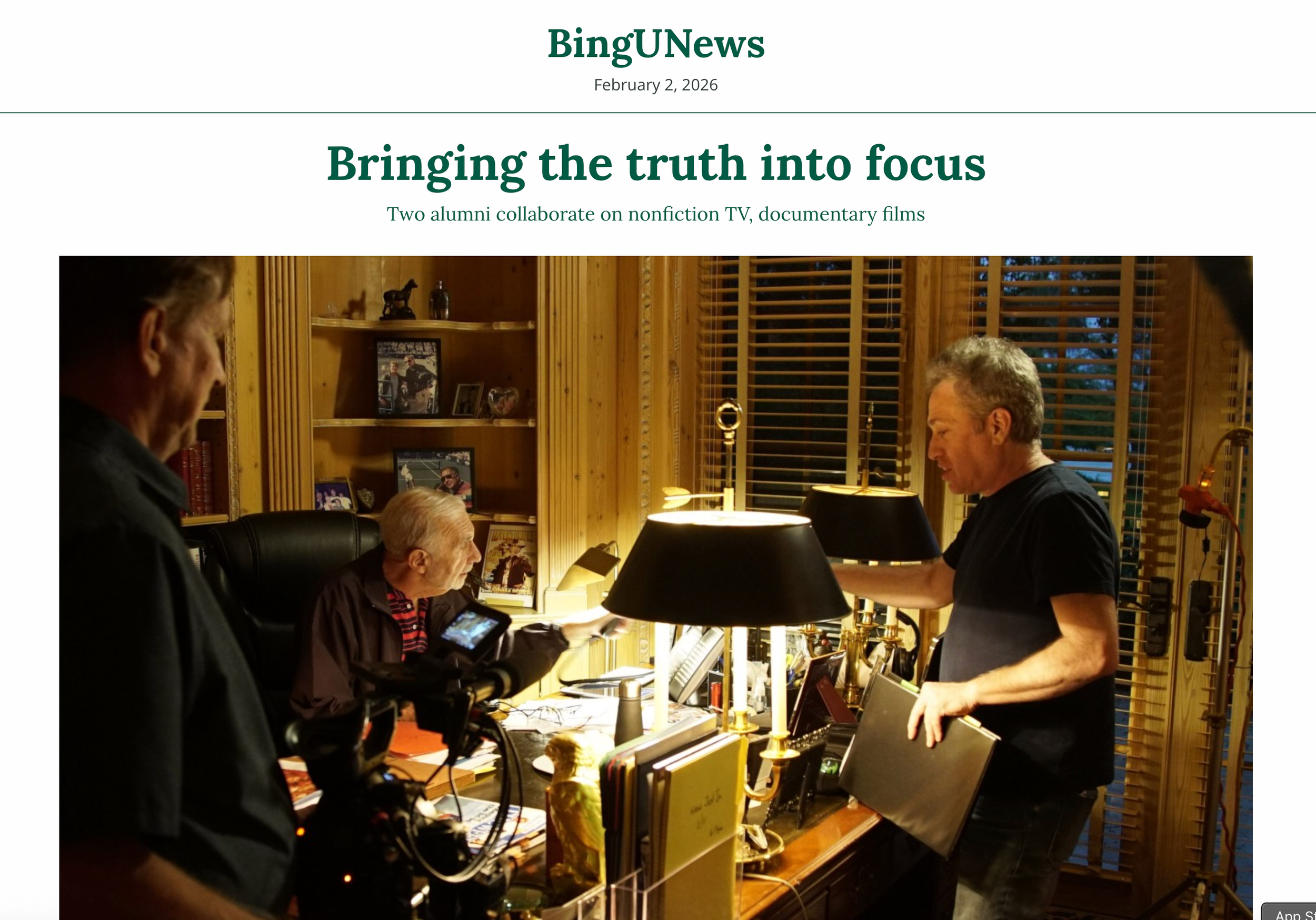 Three men in a wooden office or study, one seated at a desk with a camera, two standing, with shelves of photos and books behind them, engaged in conversation or collaboration.