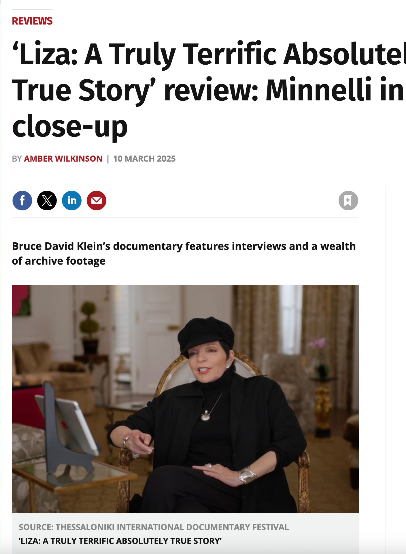 A woman in black attire and a black beret sitting in a gold ornate chair, speaking during an interview in a well-decorated room with curtains and a potted plant in the background.