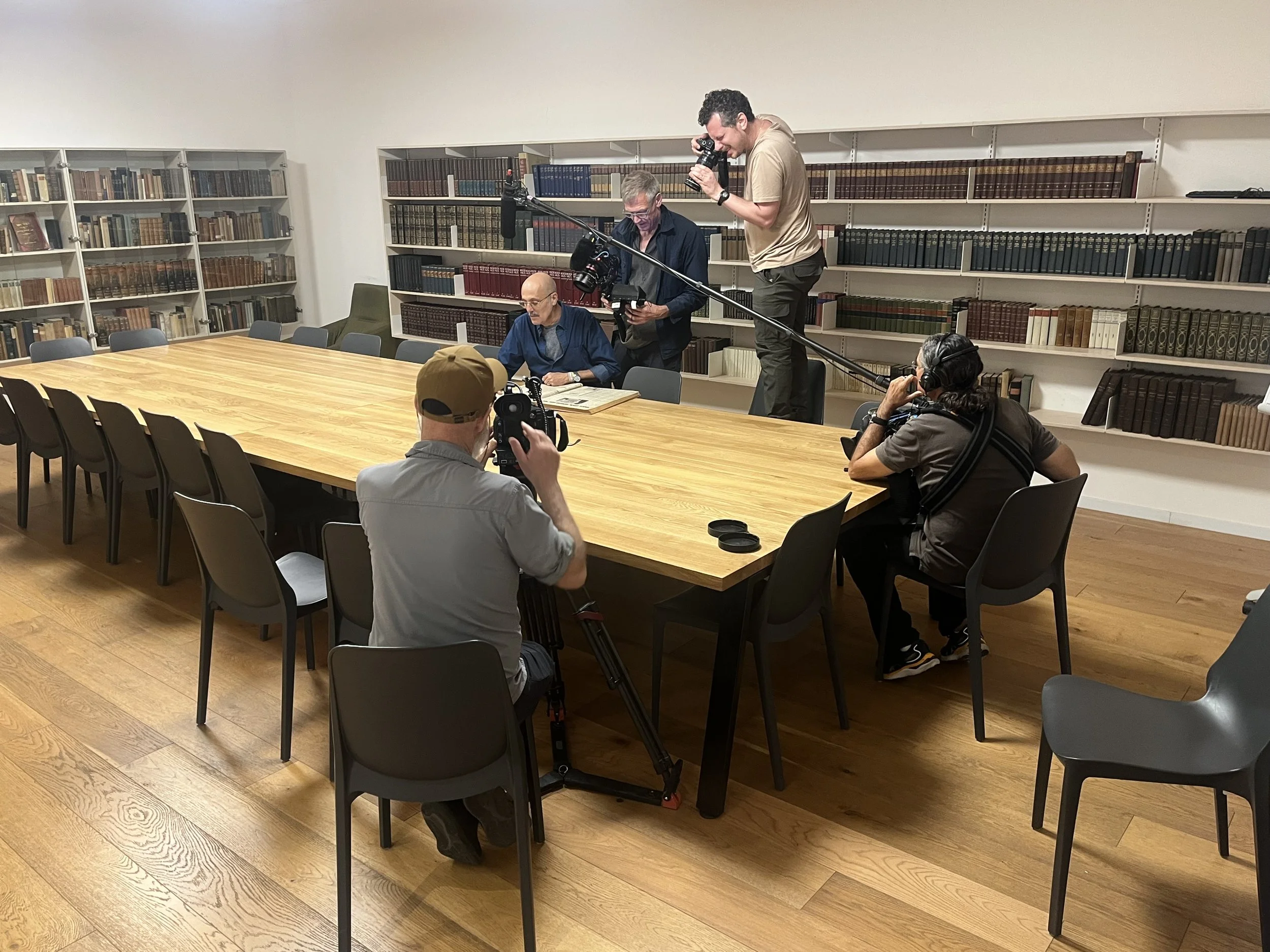 A group of five people filming a man sitting at a library table with bookshelves in the background. Some crew members are using cameras and wearing headphones.