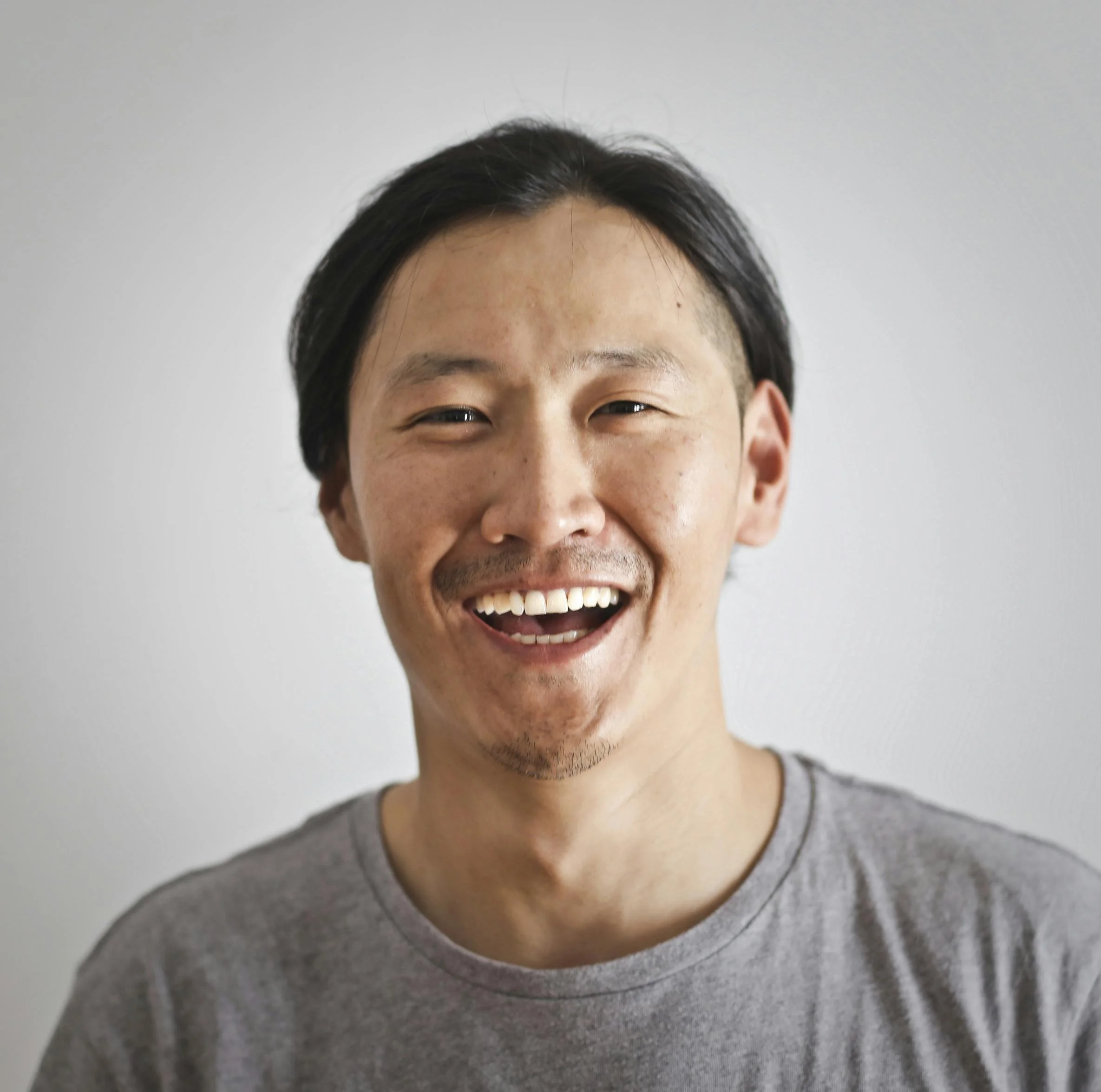 A smiling man with black hair, wearing a gray t-shirt, standing against a plain white wall.
