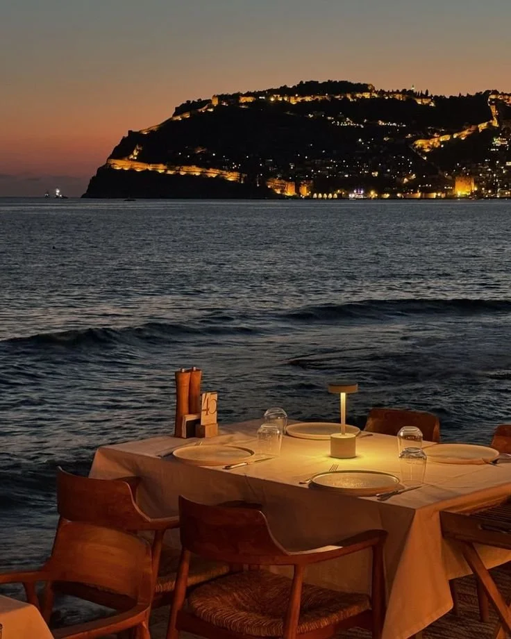 A seaside restaurant at the bosphorus in istanbul with a table set for dining, illuminated by a small lamp, overlooking the ocean at sunset with a hill or fort illuminated in the background. Symbolizing a great delicious dinner.
