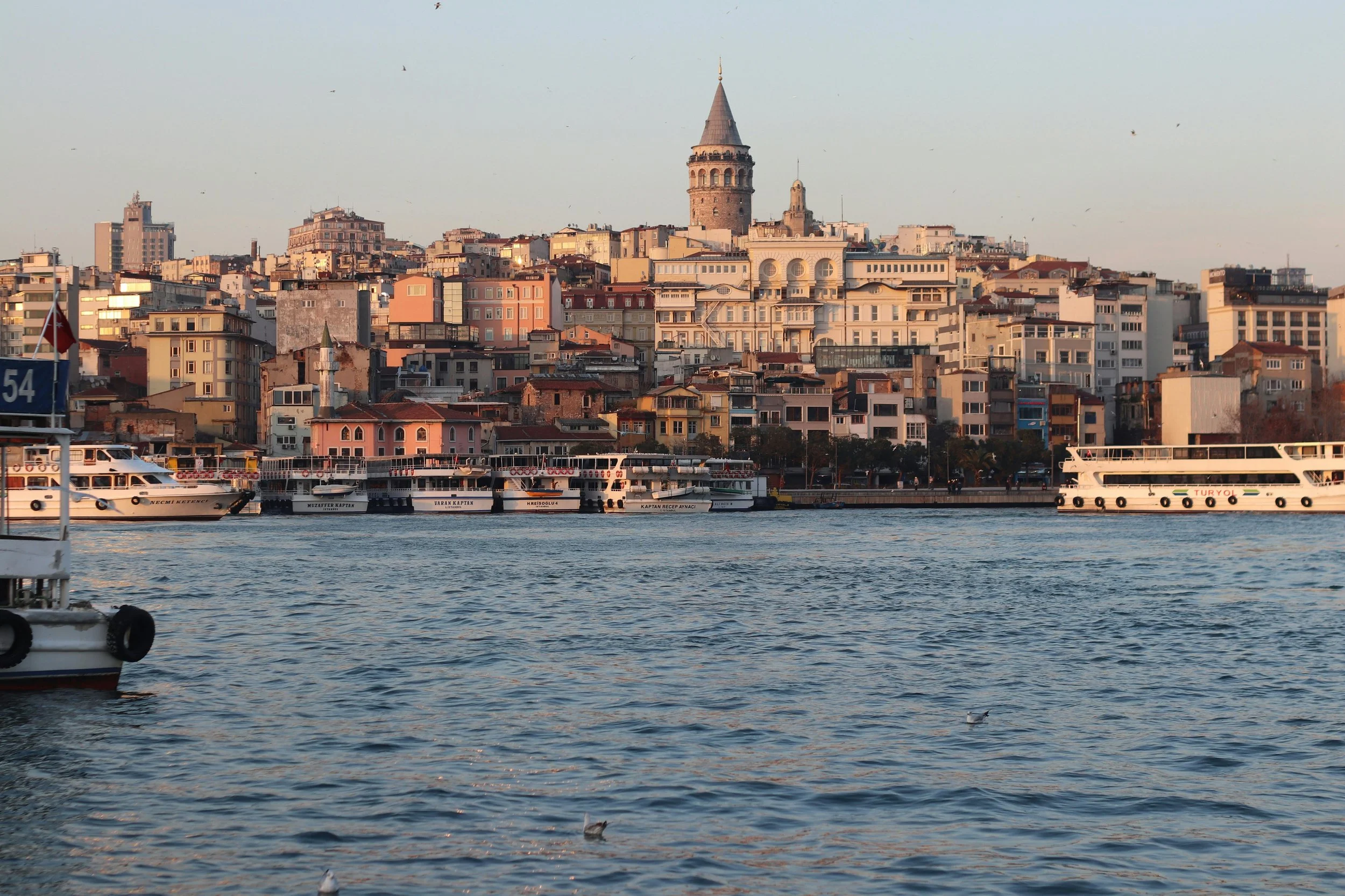 View of Istanbul skyline with historic buildings, the Galata Tower, and boats on the water during sunset.