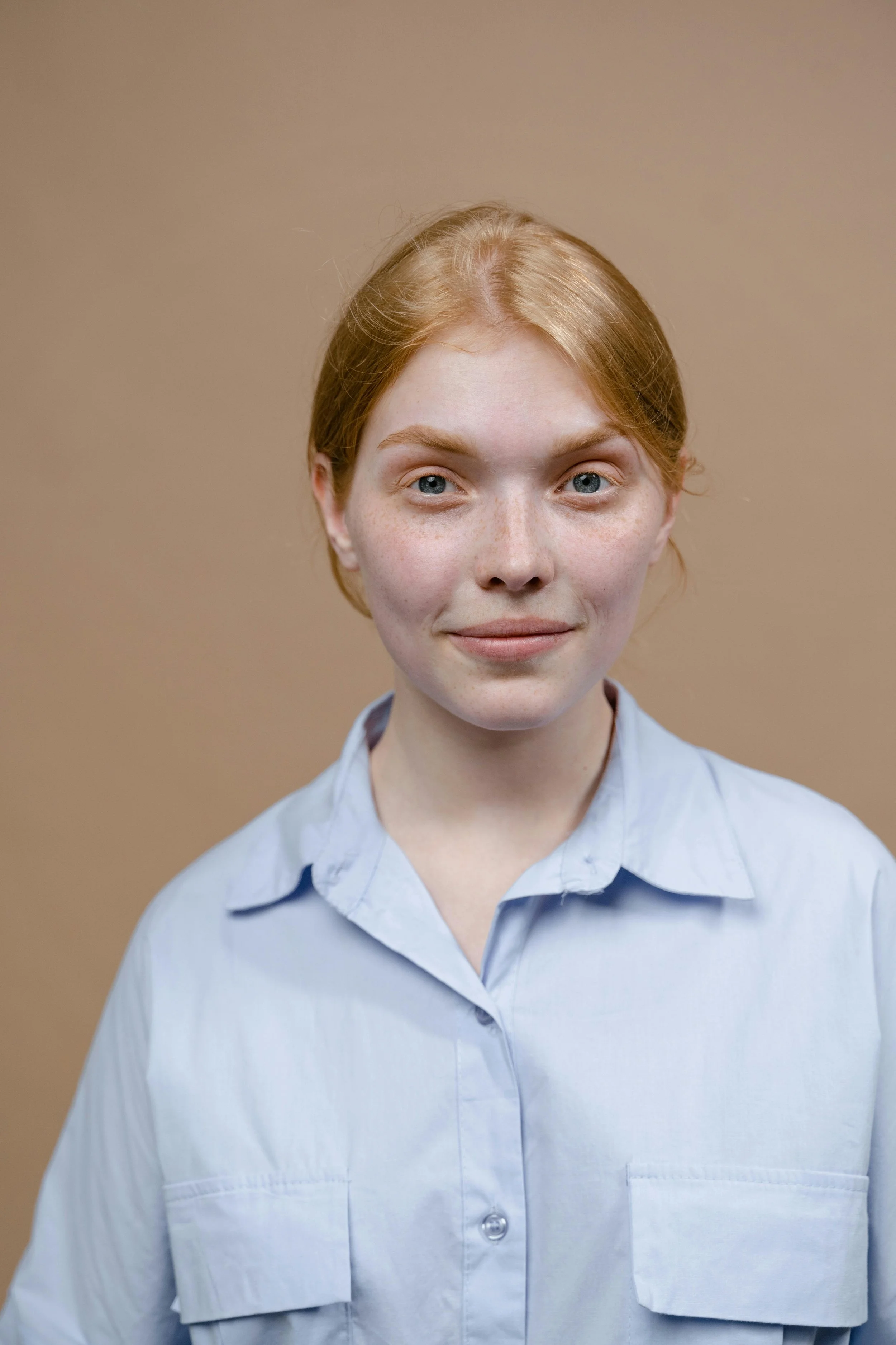 A young woman with red hair, blue eyes, and freckles, wearing a light blue button-up shirt, smiling slightly against a plain brown background.