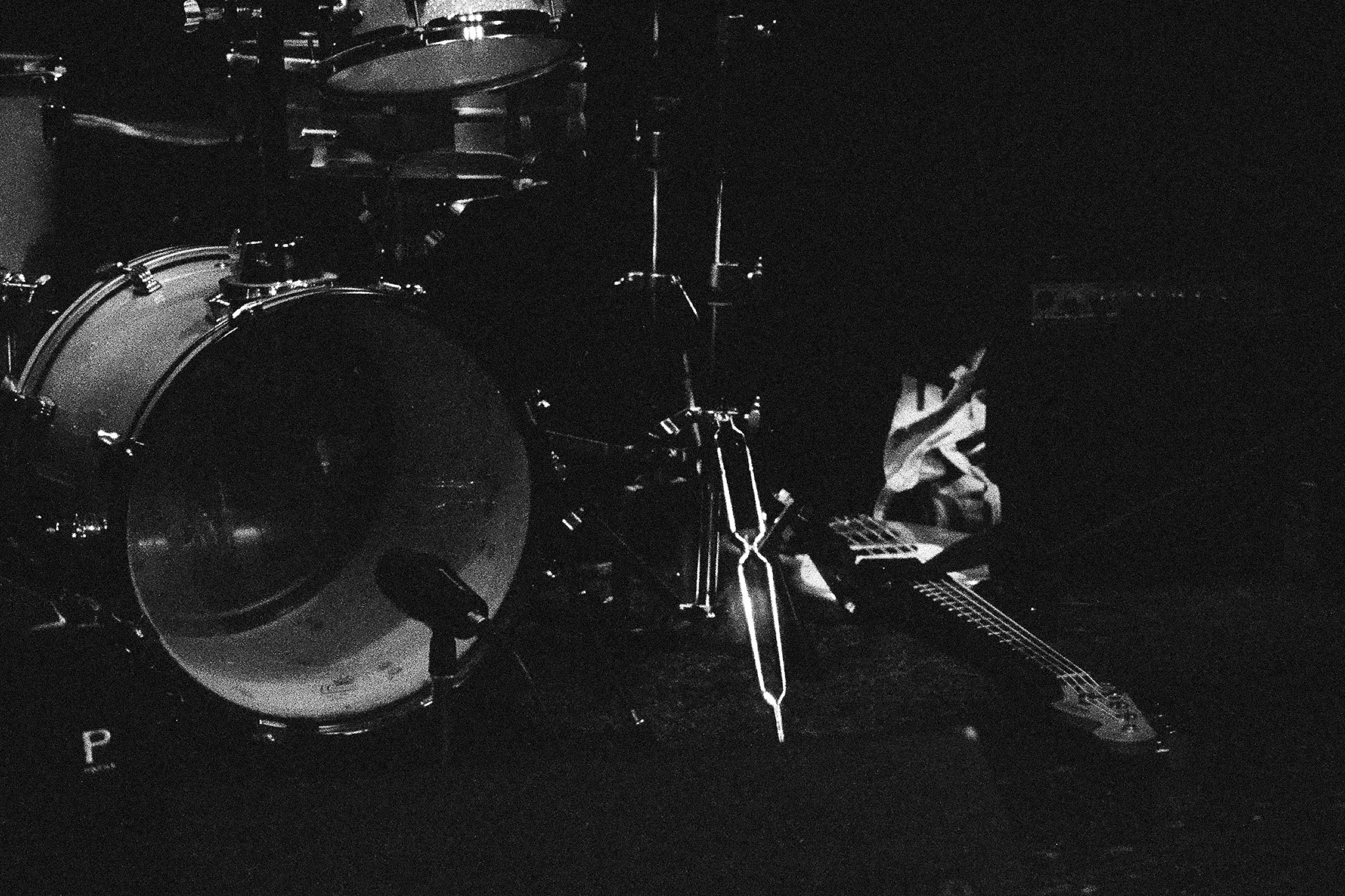 A drum set with a bass drum, snare drum, and cymbals, along with an electric guitar resting on the floor, all in a dark setting.