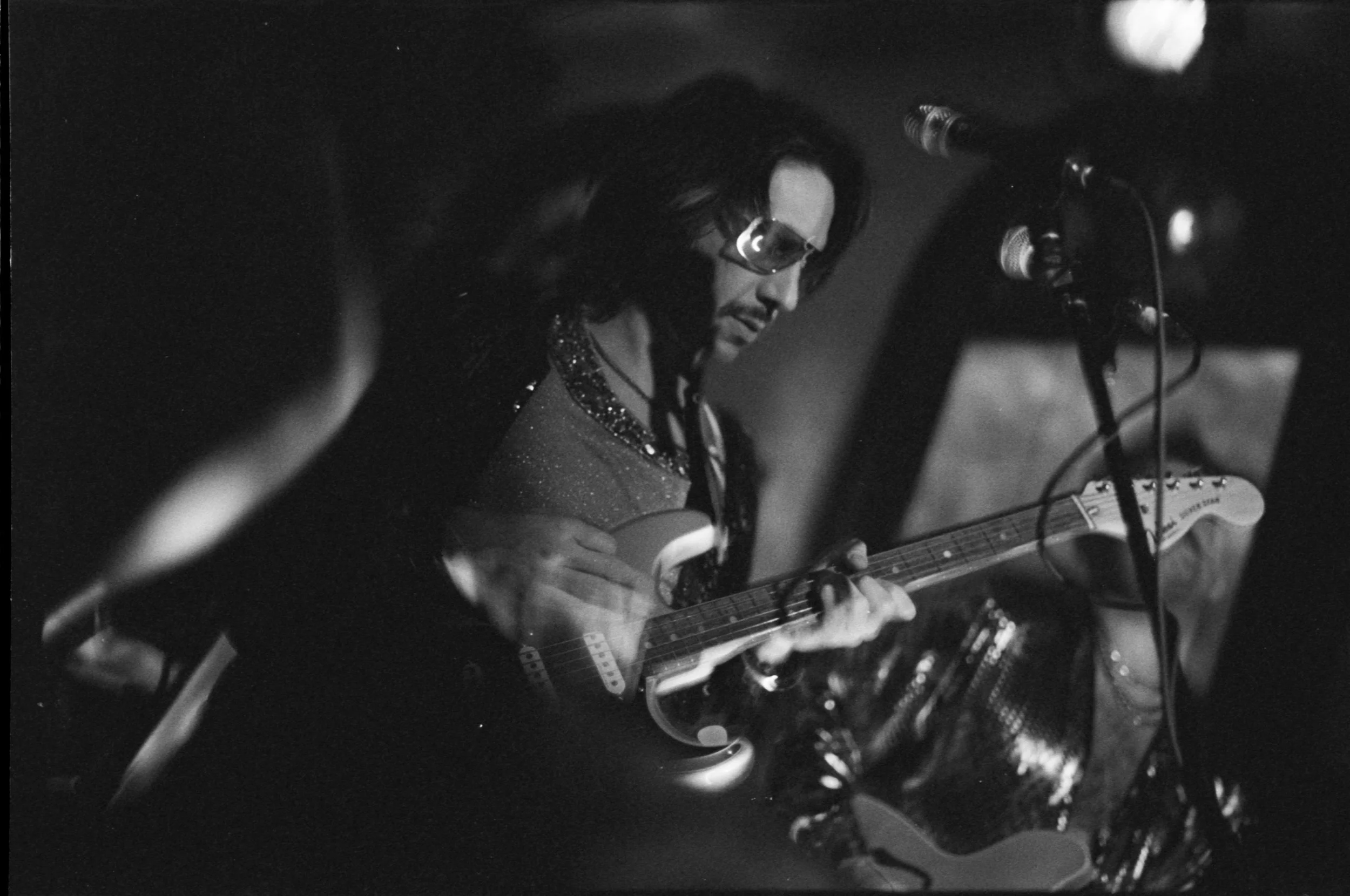 A person with long dark hair, wearing glasses and a shimmering top, playing an electric guitar on stage with microphones nearby, in a black-and-white photograph.