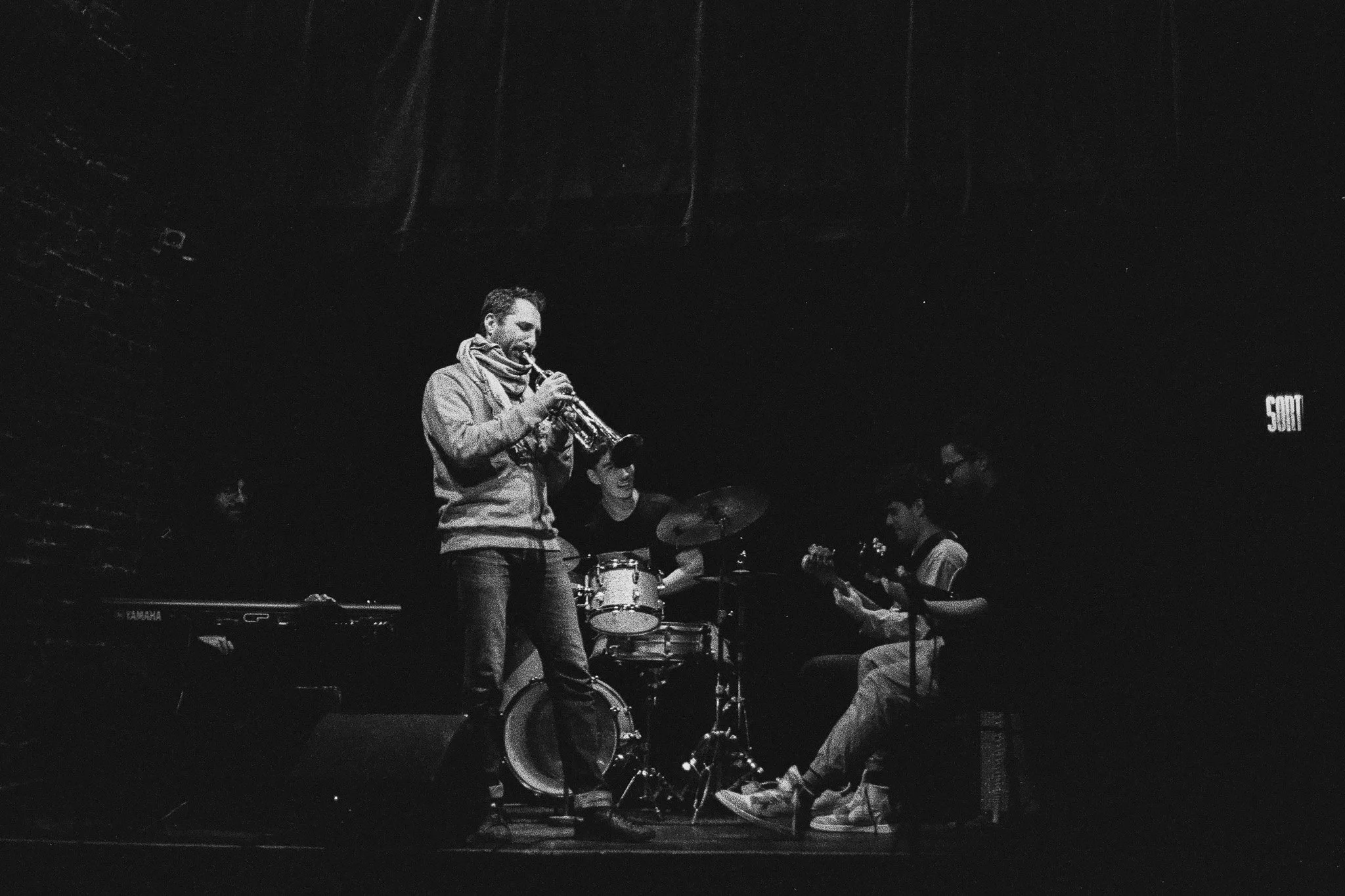 A jazz band performing on stage with a saxophonist, a drummer, and a guitarist, in a dimly lit venue.