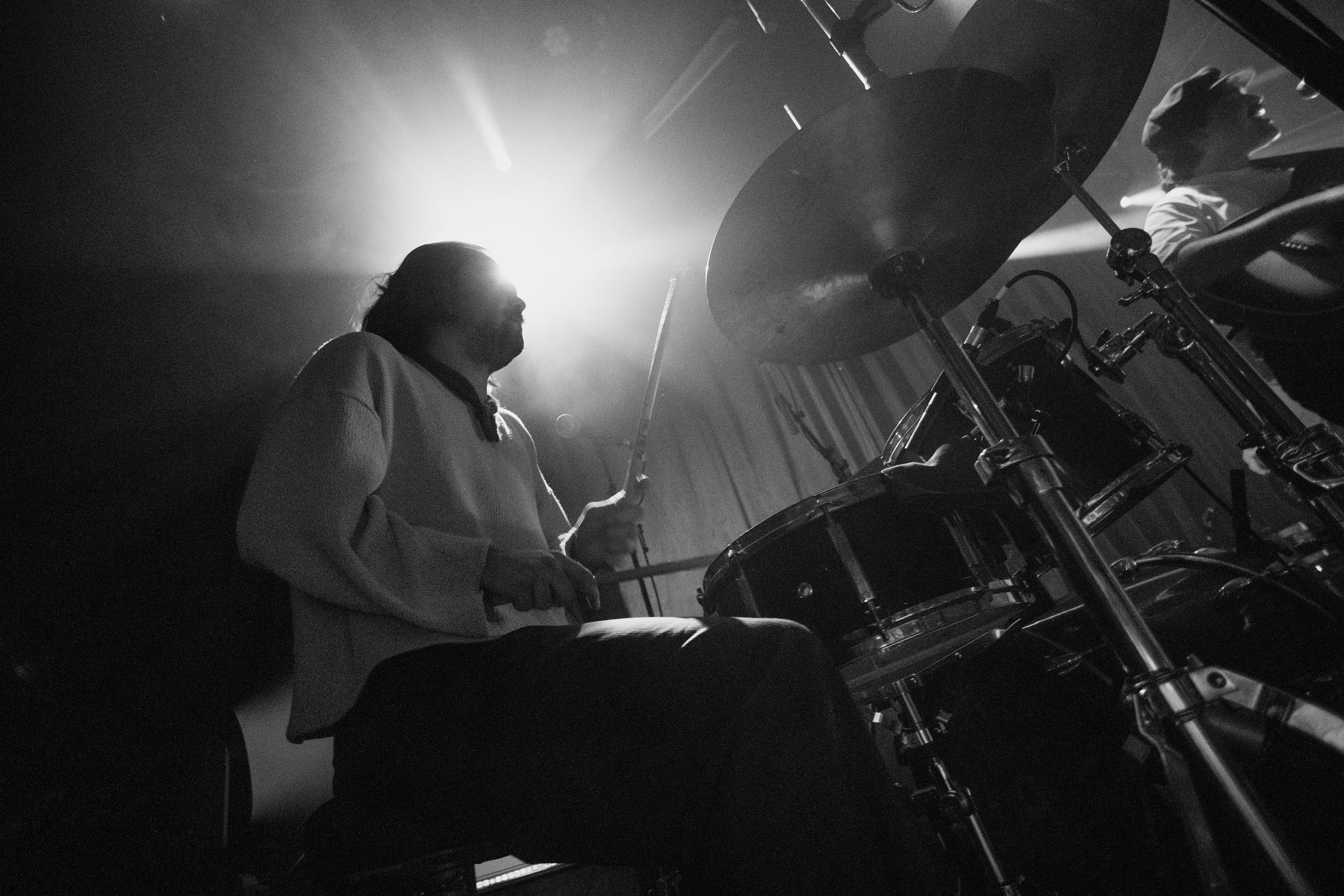 A person playing drums on stage with backlighting creating a silhouette, with another musician slightly visible in the background.
