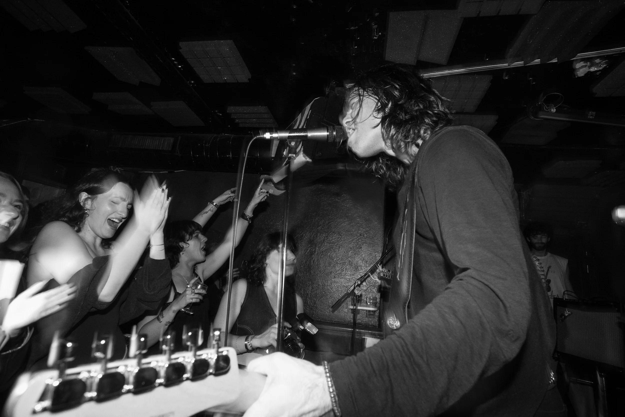 A black and white photo of a live music performance where a musician is singing into a microphone, and the audience is clapping and cheering.