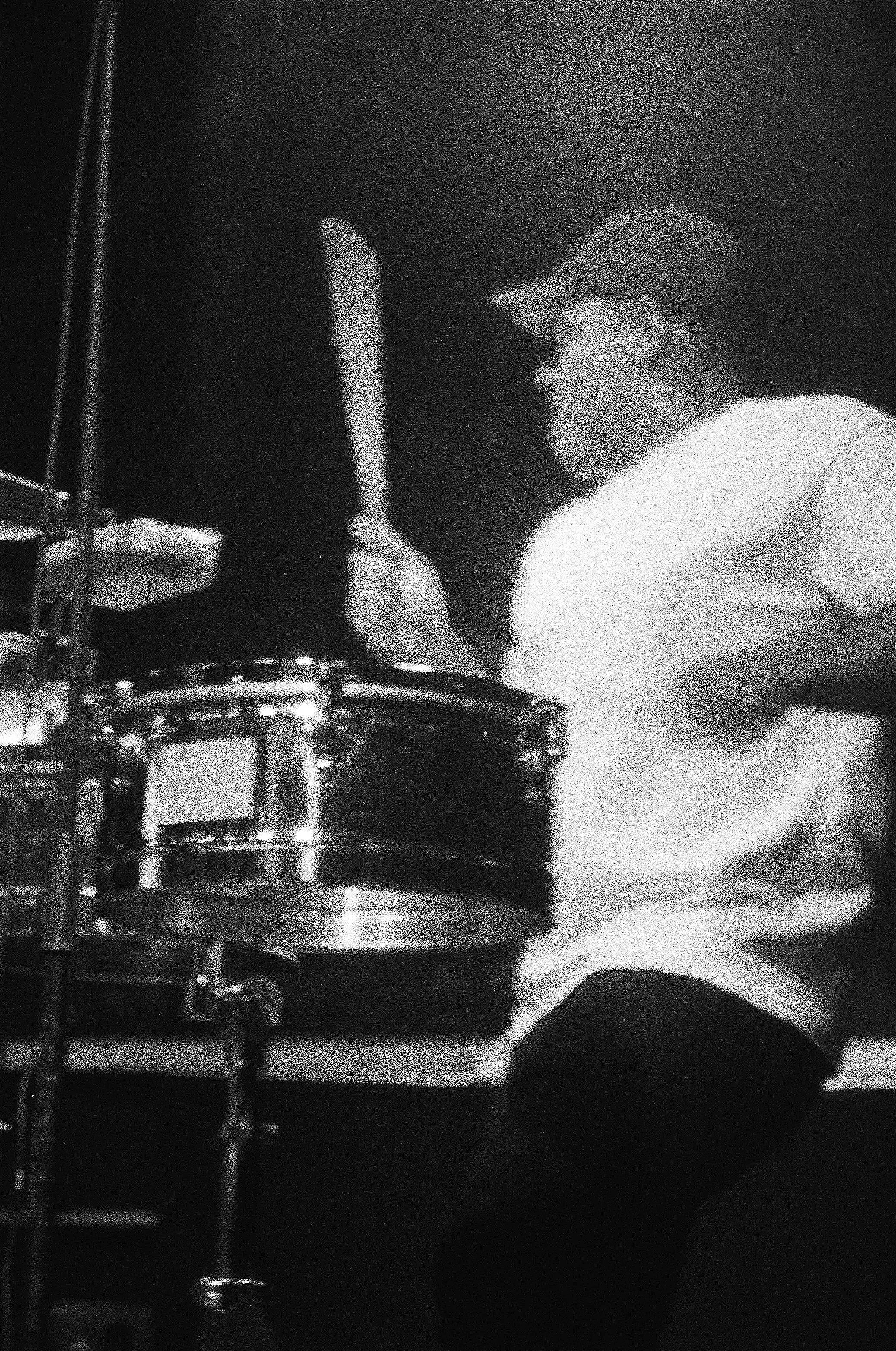 A person wearing glasses, a cap, and a white shirt playing a drum set in black and white.