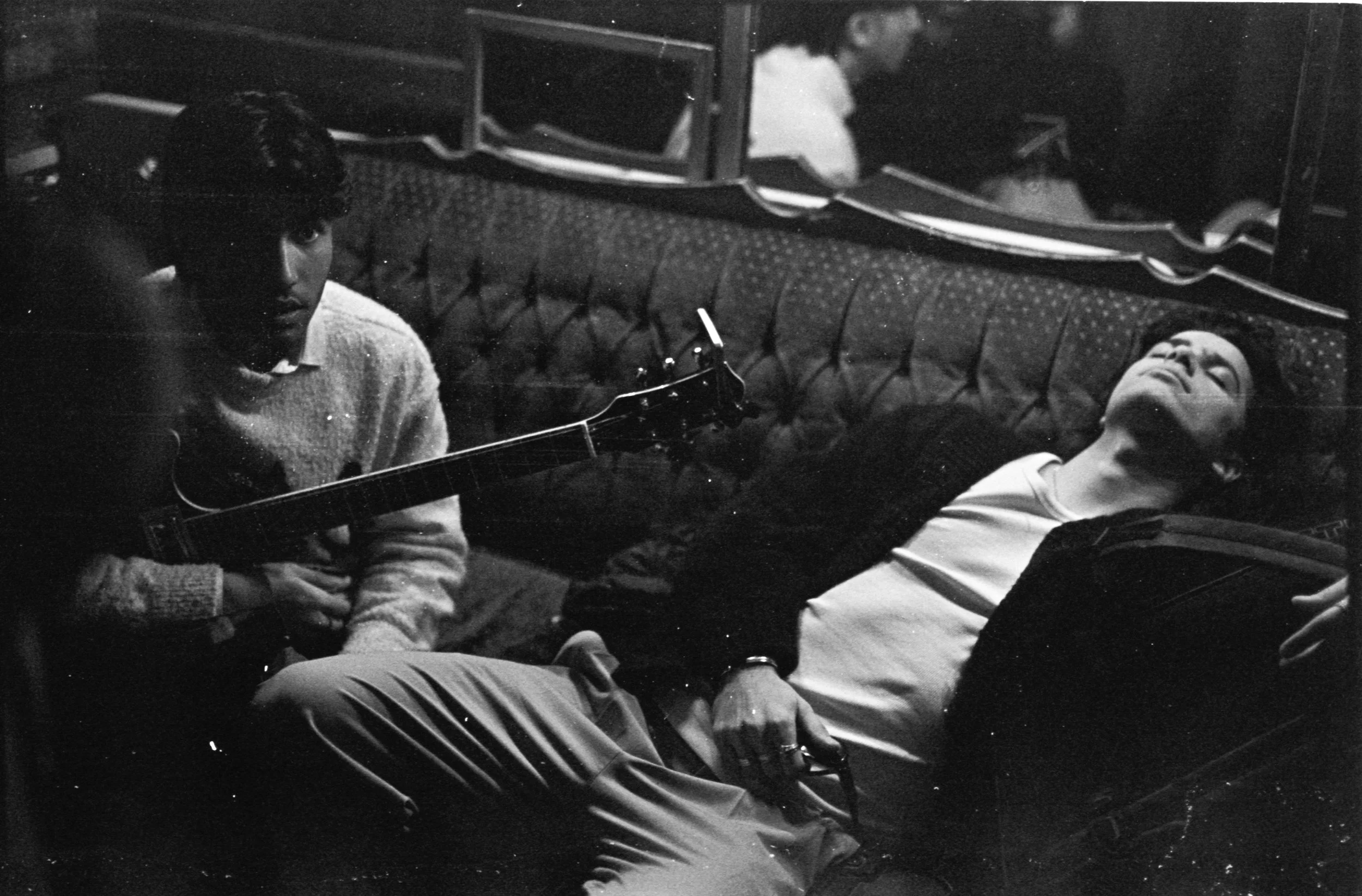 Two young men in a dimly lit room, one sitting with a guitar and the other lying on a couch, eyes closed, with a mirror reflecting their images.