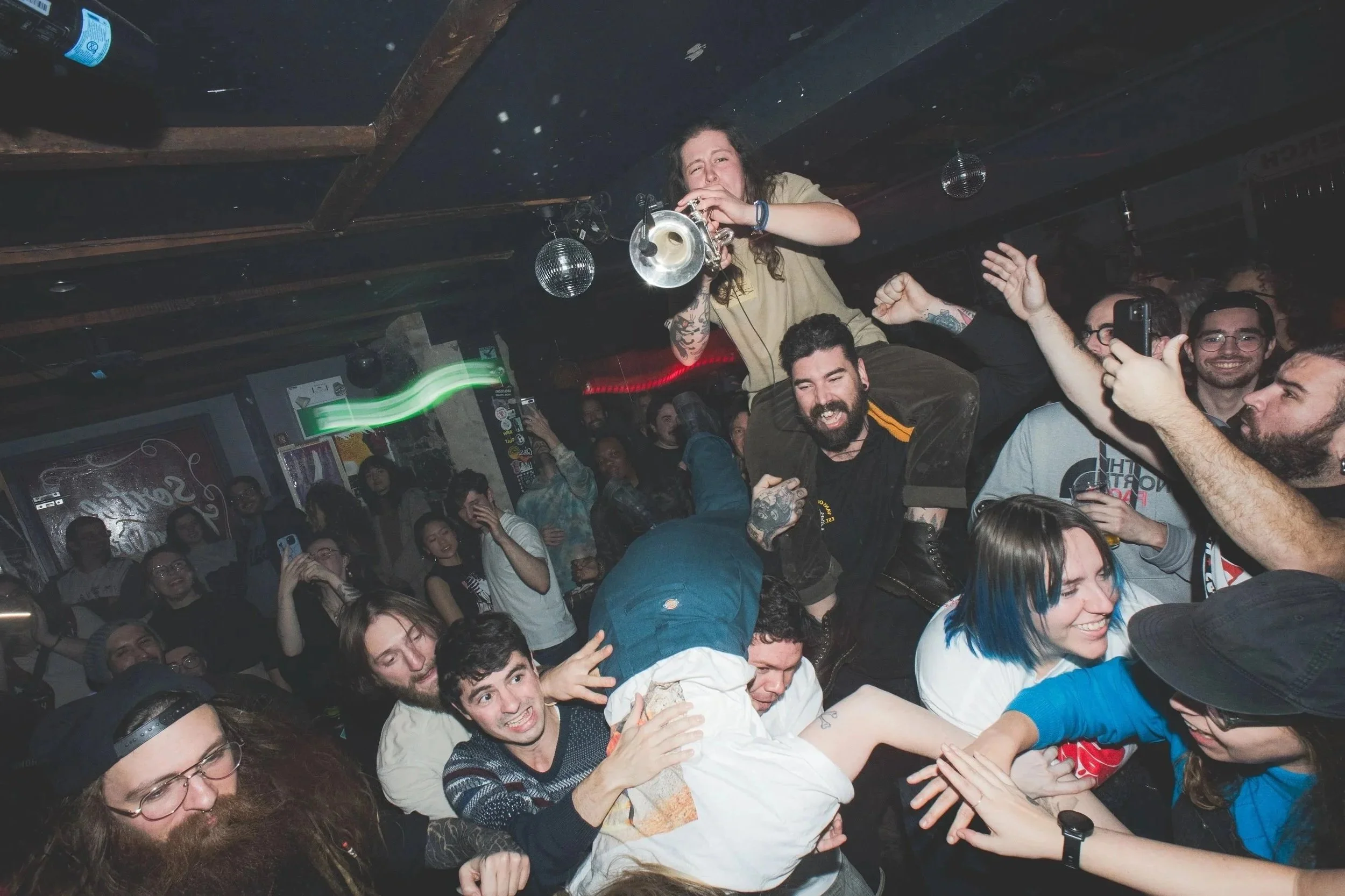 People at a lively party, some crowd-surfing and taking photos, in a dimly lit indoor venue with disco balls overhead.