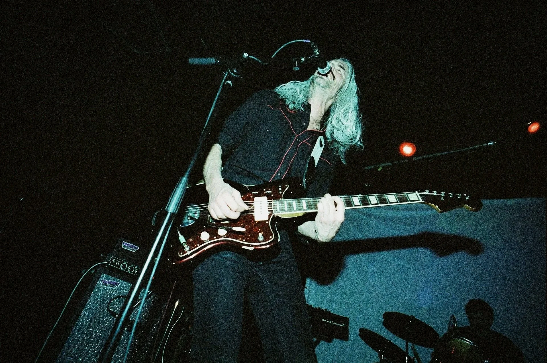 A musician with long gray hair singing into a microphone and playing an electric guitar on stage in a dark setting.
